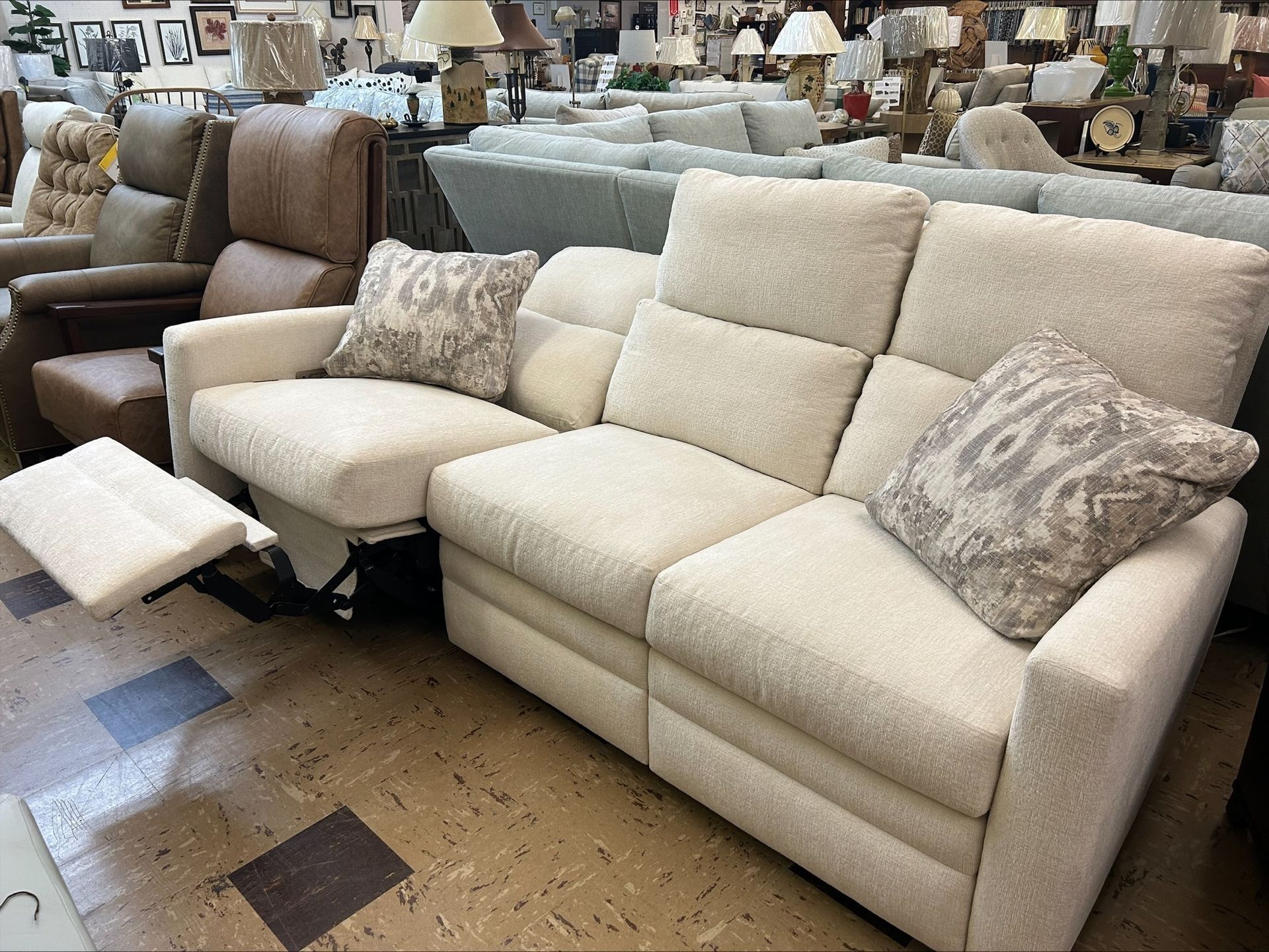 Beige reclining sofa in a furniture store, with one footrest extended, patterned pillows, and other furniture visible.