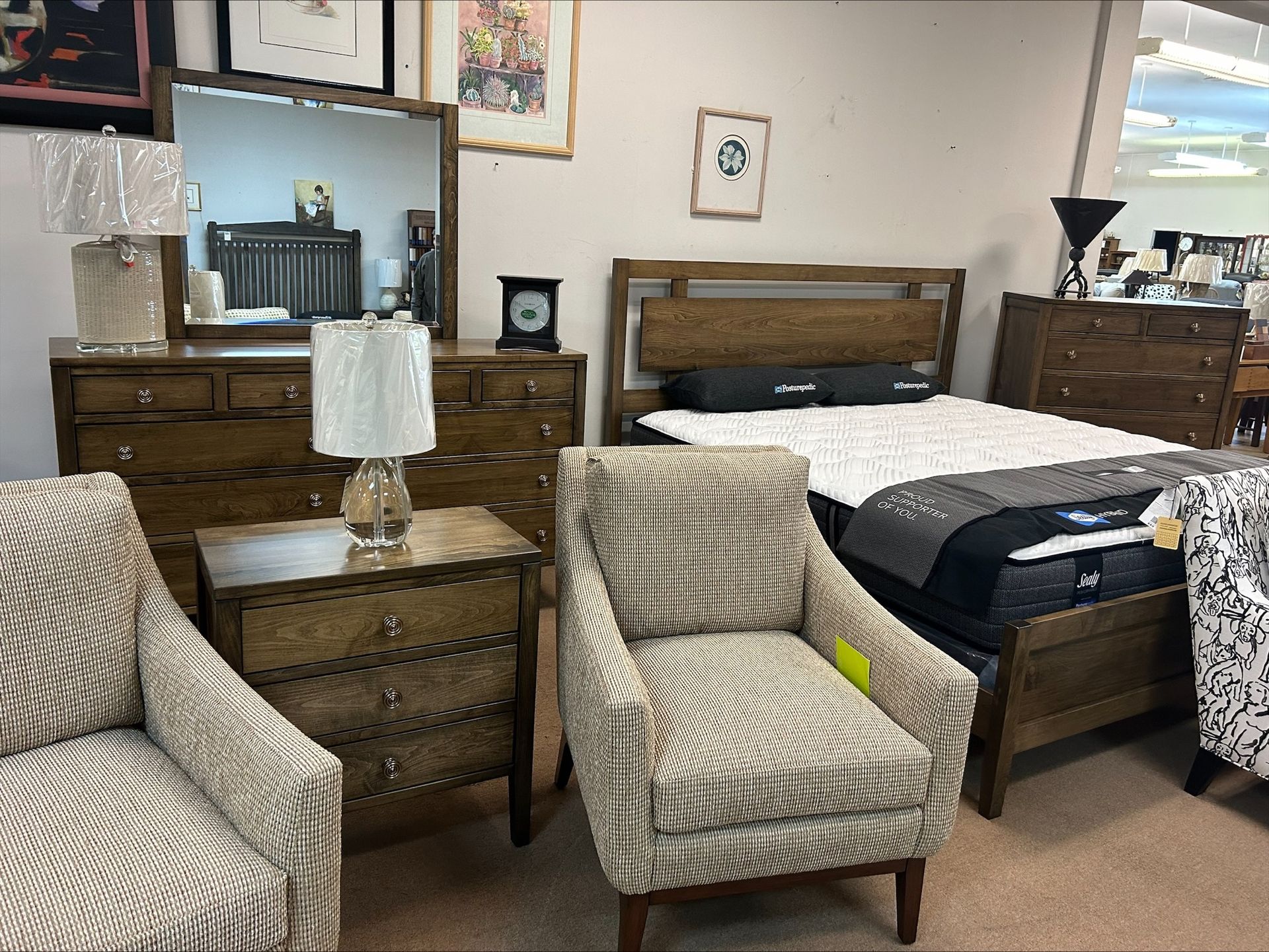 Bedroom furniture set in a showroom, including a bed, dresser, nightstand, chairs, and artwork on the walls.