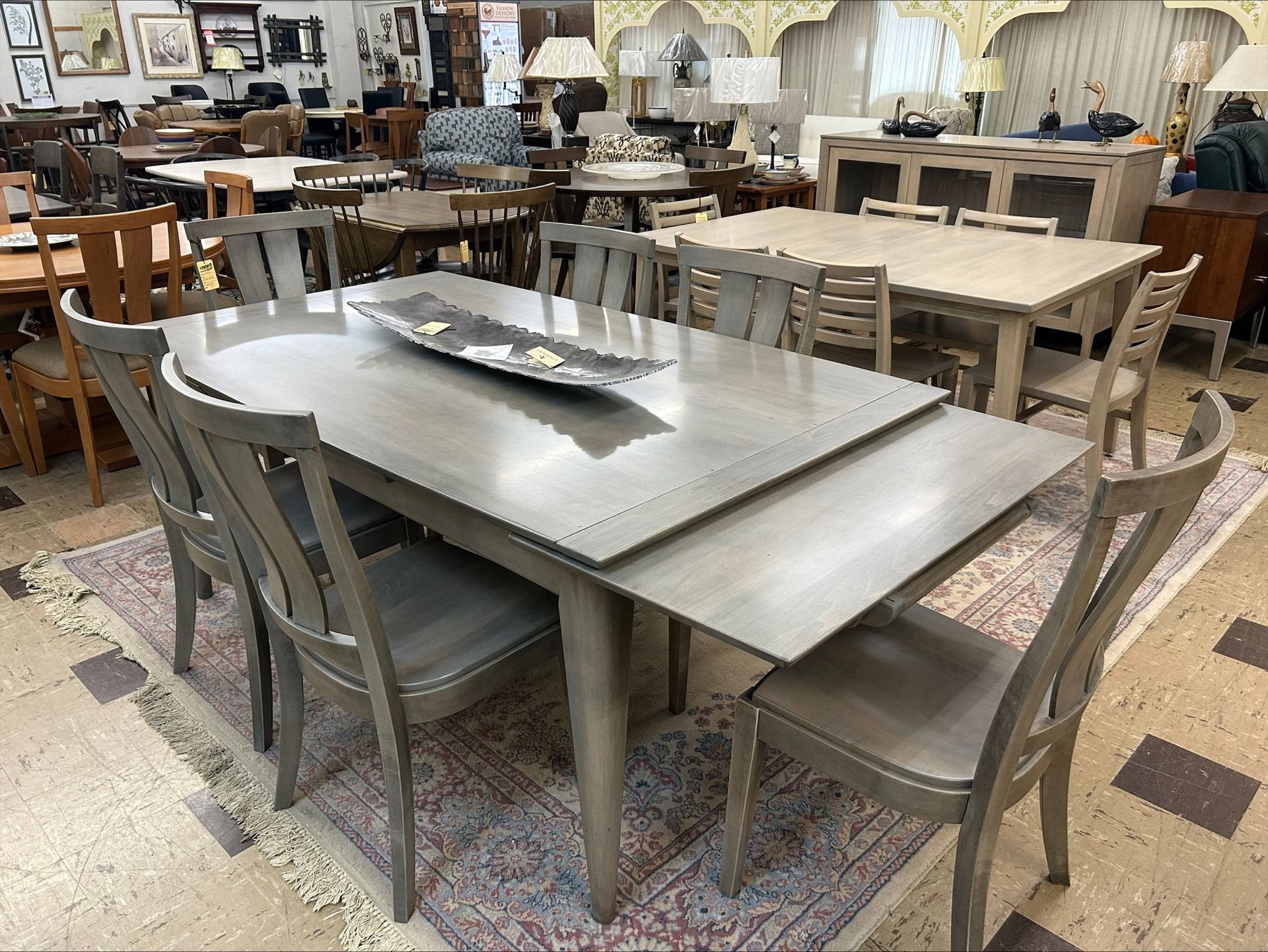Gray dining table with chairs in a furniture store, set on a patterned rug.