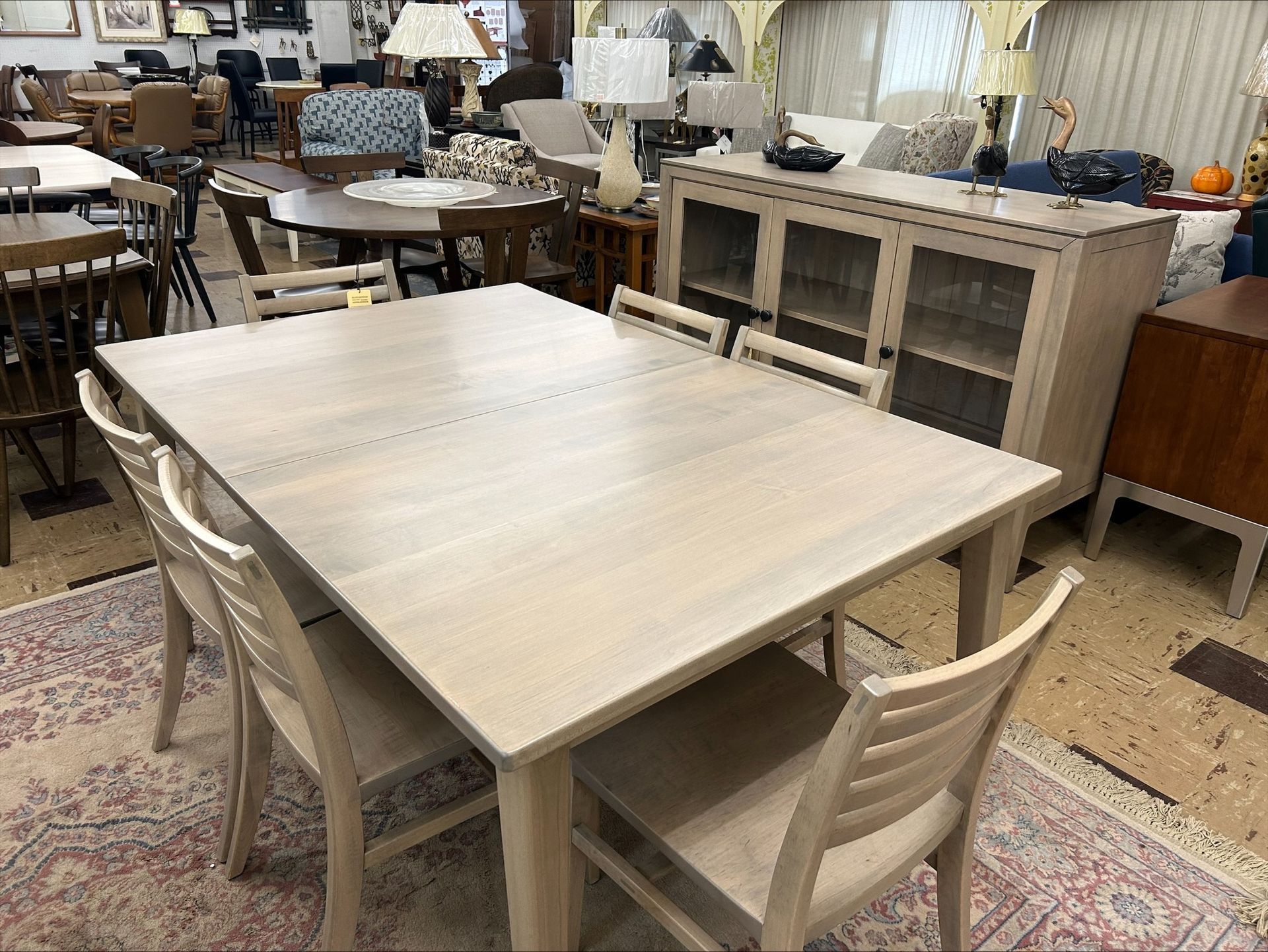 Dining table with four chairs, light wood color, on a patterned rug in a furniture store.
