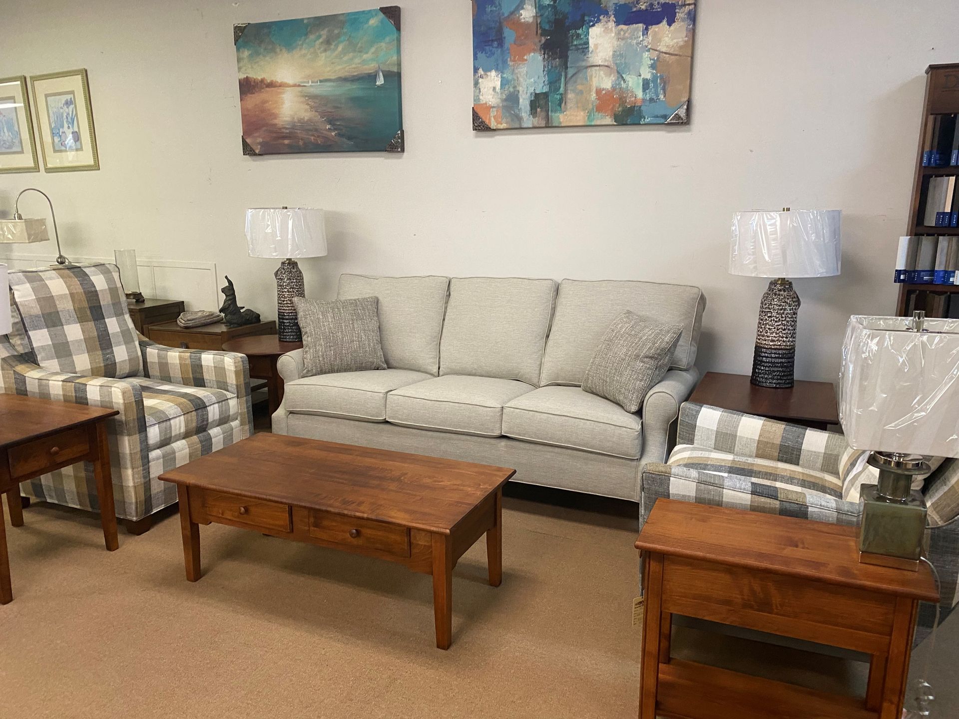 Living room furniture display: sofa, armchairs, coffee table, lamps, paintings, and shelving unit.