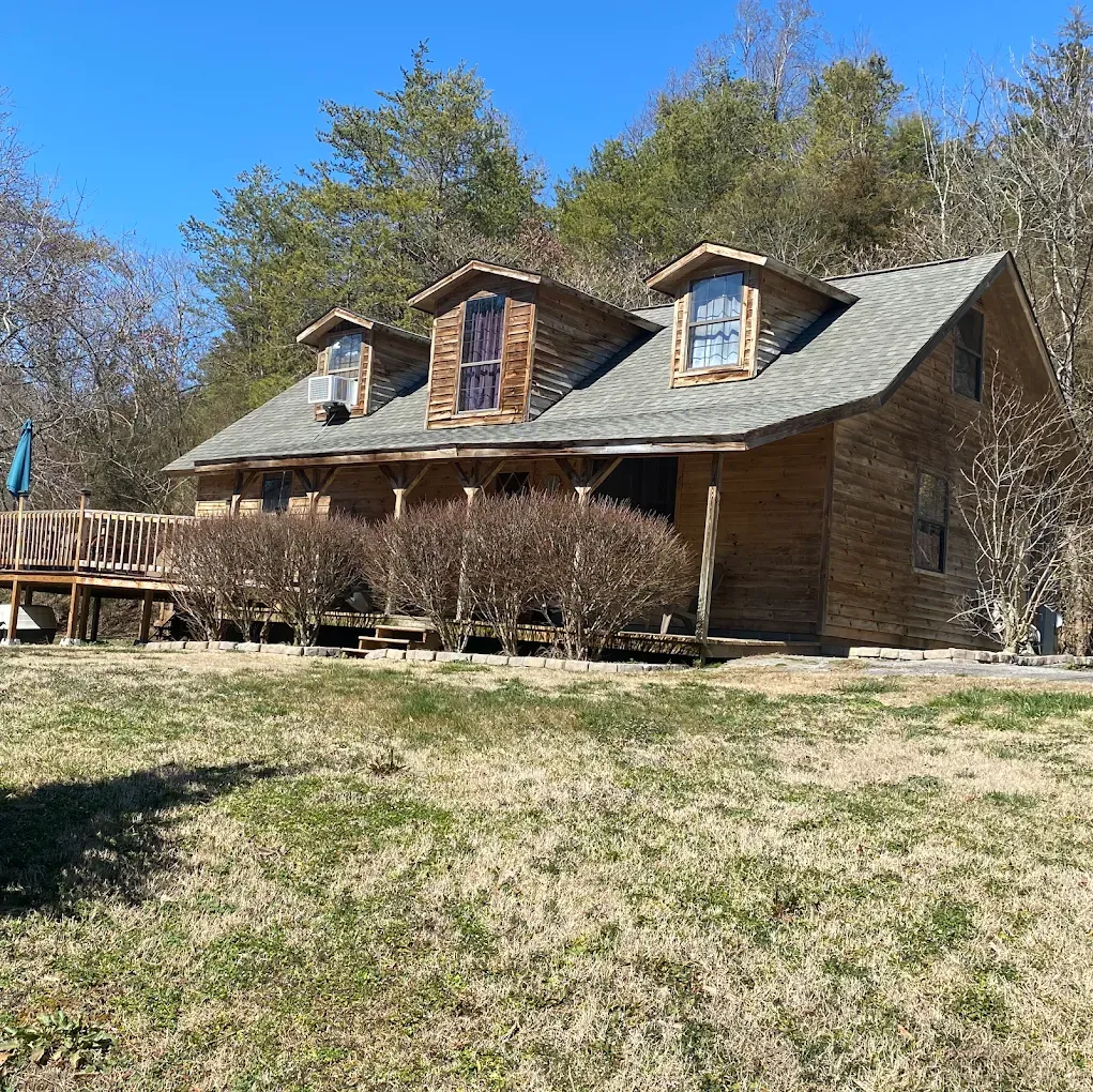 Log cabin with three dormers, a deck, and a yard, surrounded by trees on a sunny day.