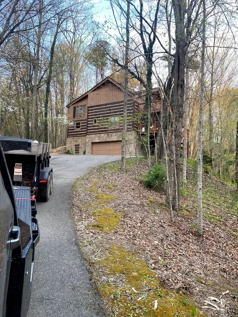 A brown, two-story house on a hillside with a long driveway; a vehicle with a trailer is parked on the road.