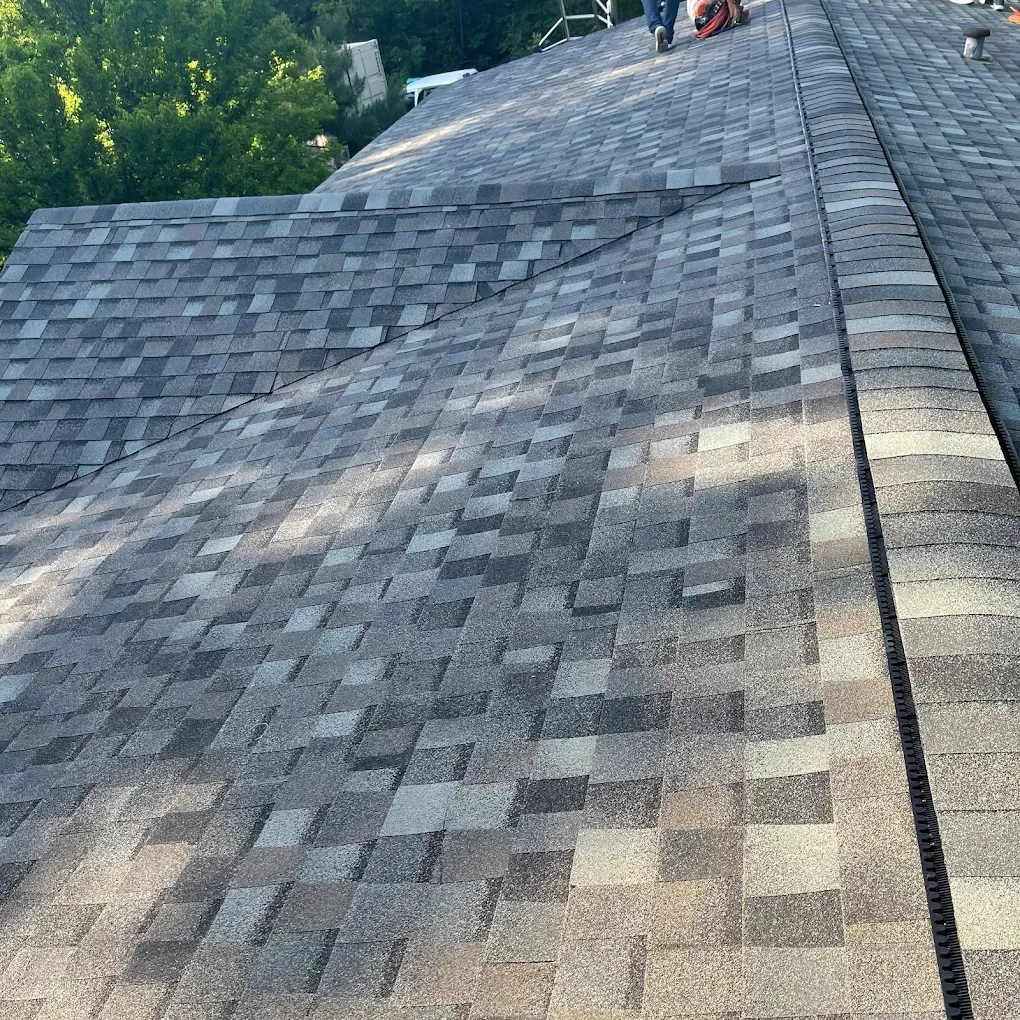 Gray asphalt shingle roof with a person working, surrounded by trees.