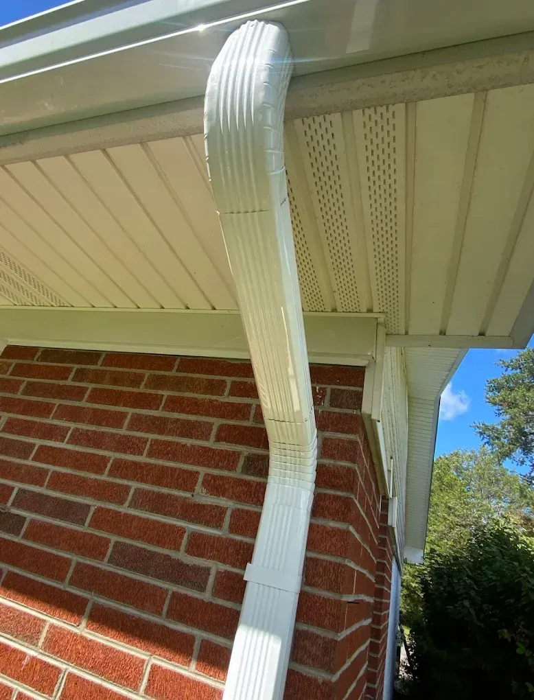 White gutter attached to a brick building's edge with white trim and siding, a blue sky visible.