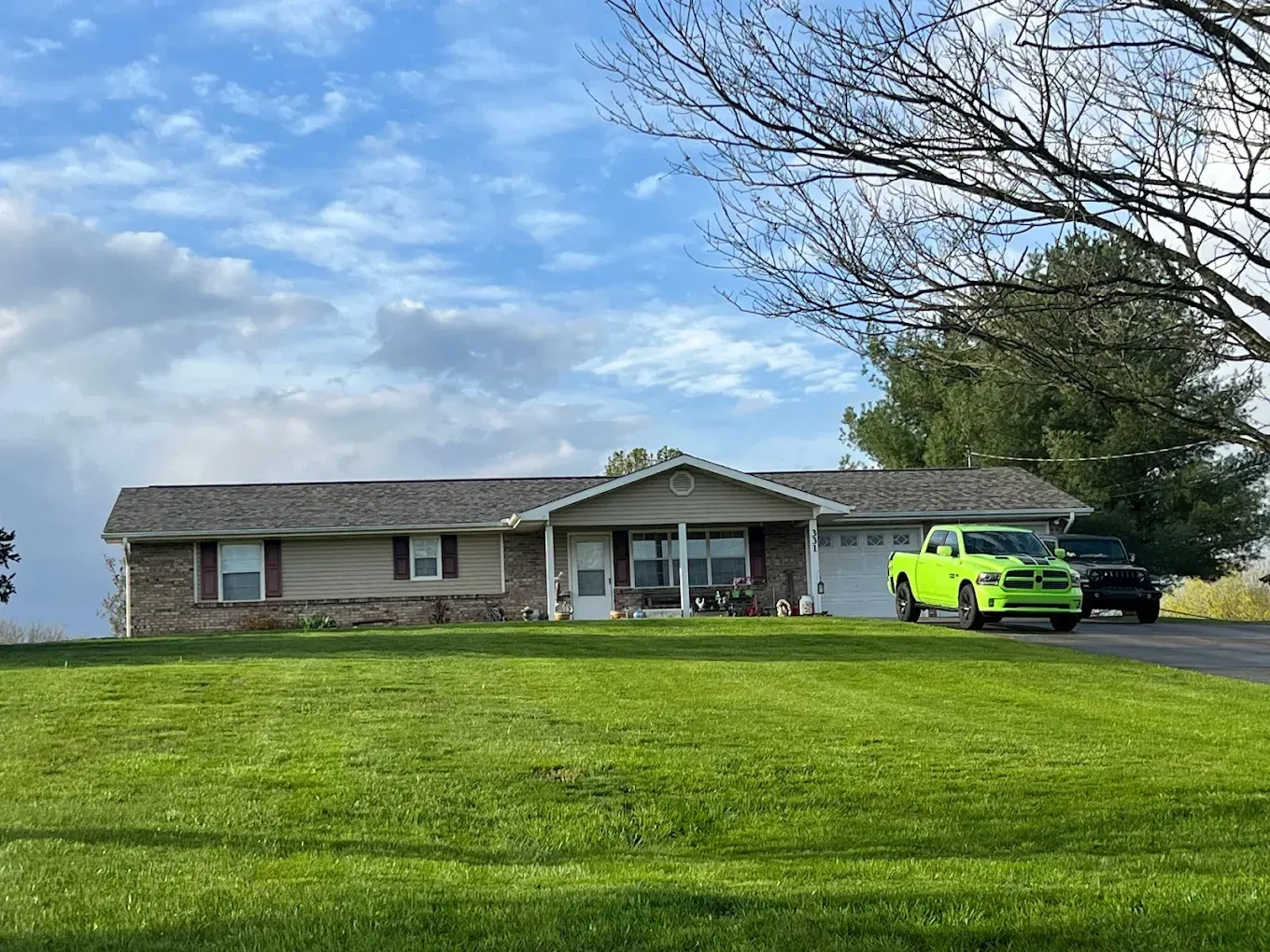 Ranch-style house with green lawn; a green truck and black vehicle are parked in front on a cloudy day.