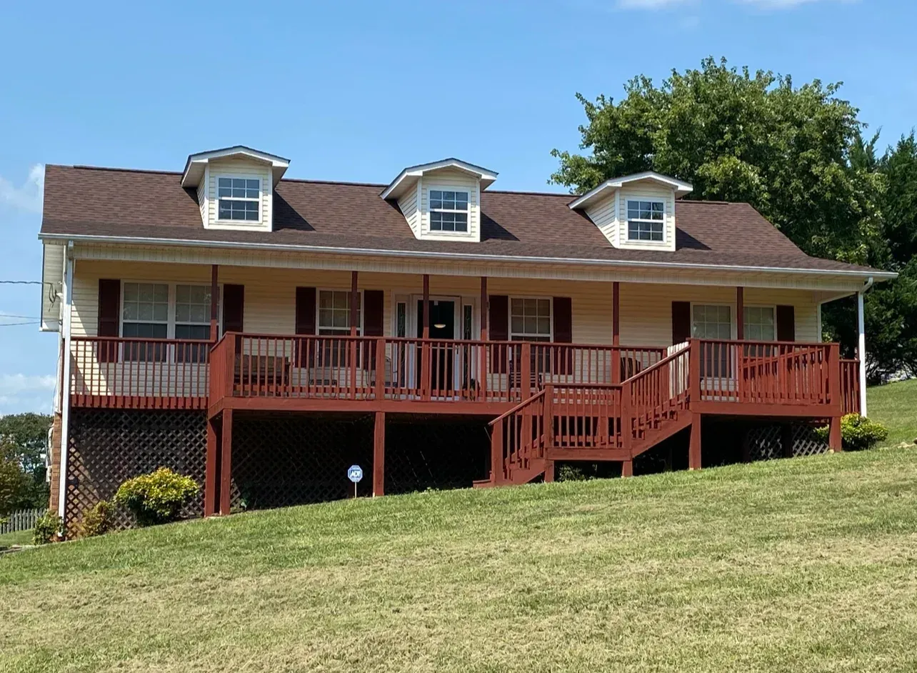 Yellow house with brown roof, deck, and shutters, on a grassy hill under a blue sky.