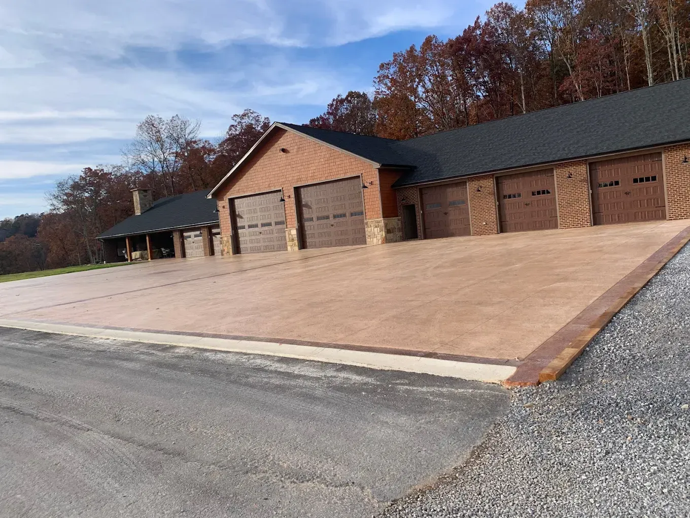 Brick garage with multiple brown doors and a large, light-brown concrete driveway. Autumn trees in the background.