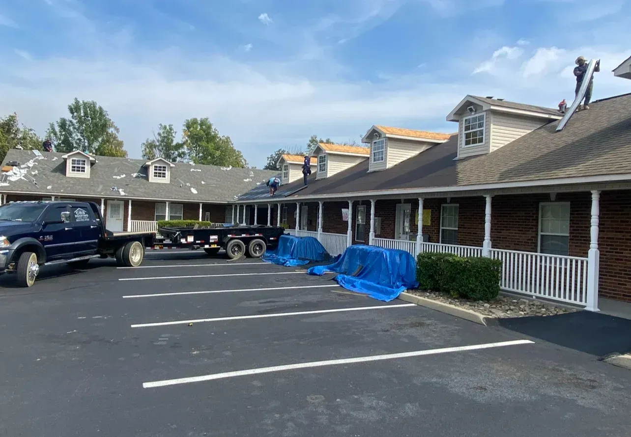 Roofing work in progress on a single-story brick building. Blue tarps cover items below. A truck and trailer are present.