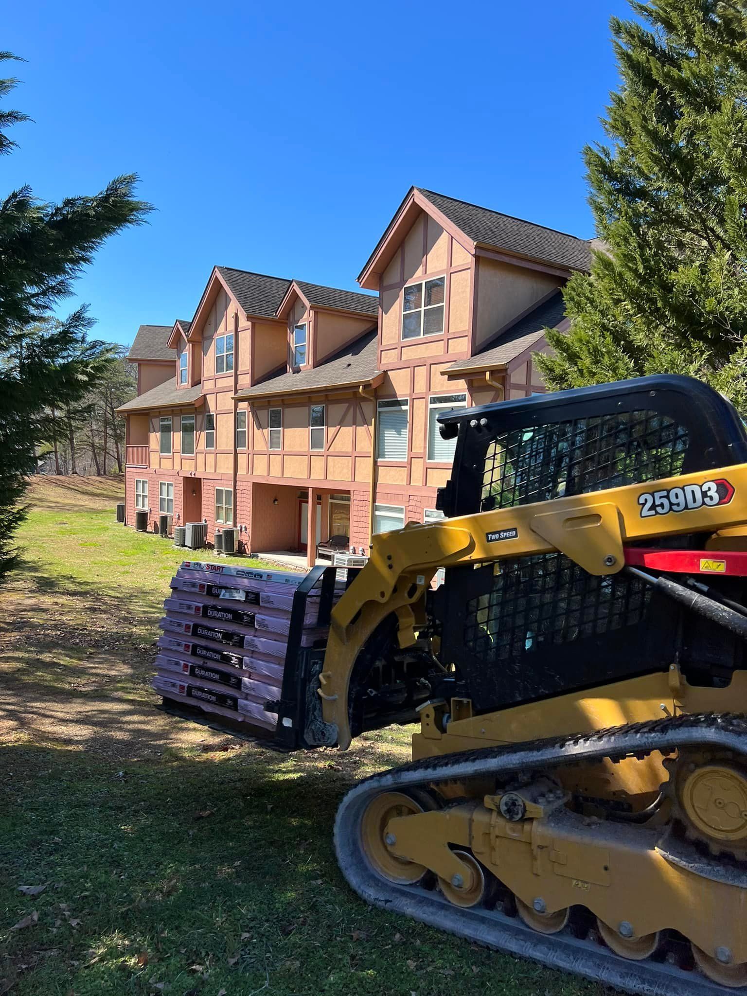 A yellow skid steer carrying purple insulation near a row of tan townhouses on a sunny day.