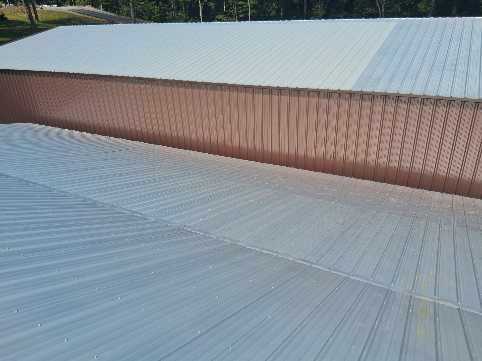An angled, high-angle view of a metal roof with light-colored corrugated panels below a brown vertical-sided wall.