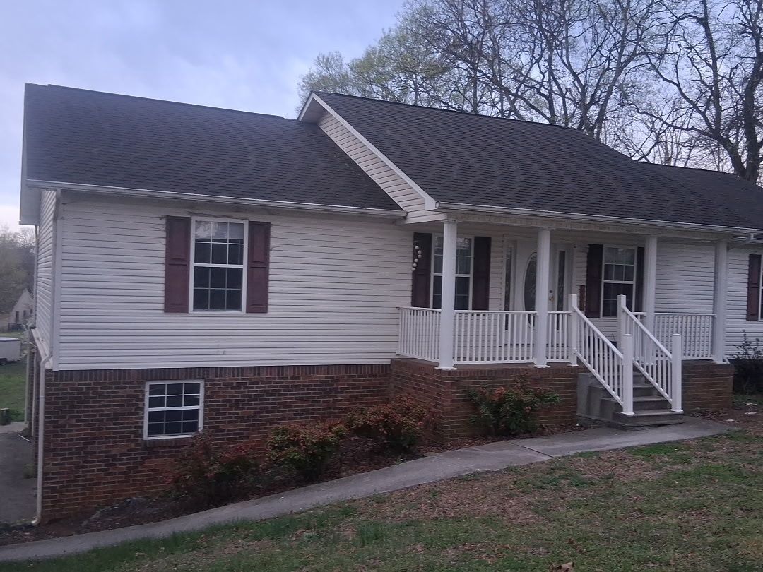 A white, single-story house with a brick foundation, dark shingled roof, and a front porch with white railings.