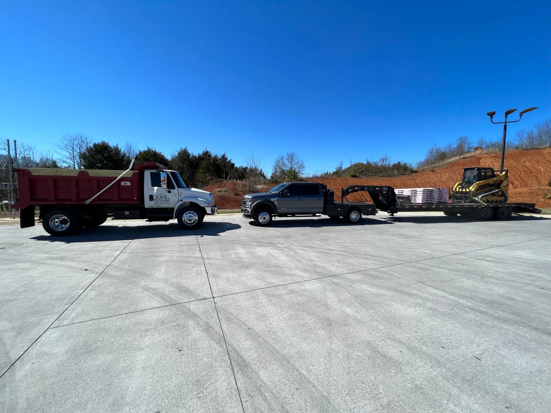 A red dump truck, a dark pickup truck, and a trailer carrying construction equipment parked on a large, paved lot.