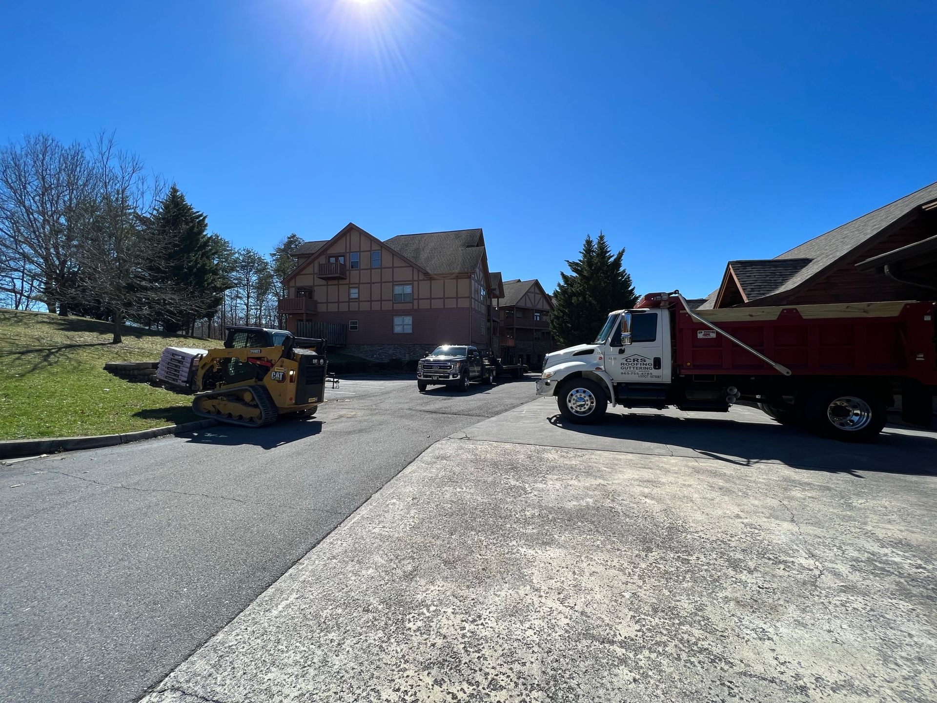 A construction site with a yellow skid steer, a white dump truck, and a pickup truck parked near a building under a sun.
