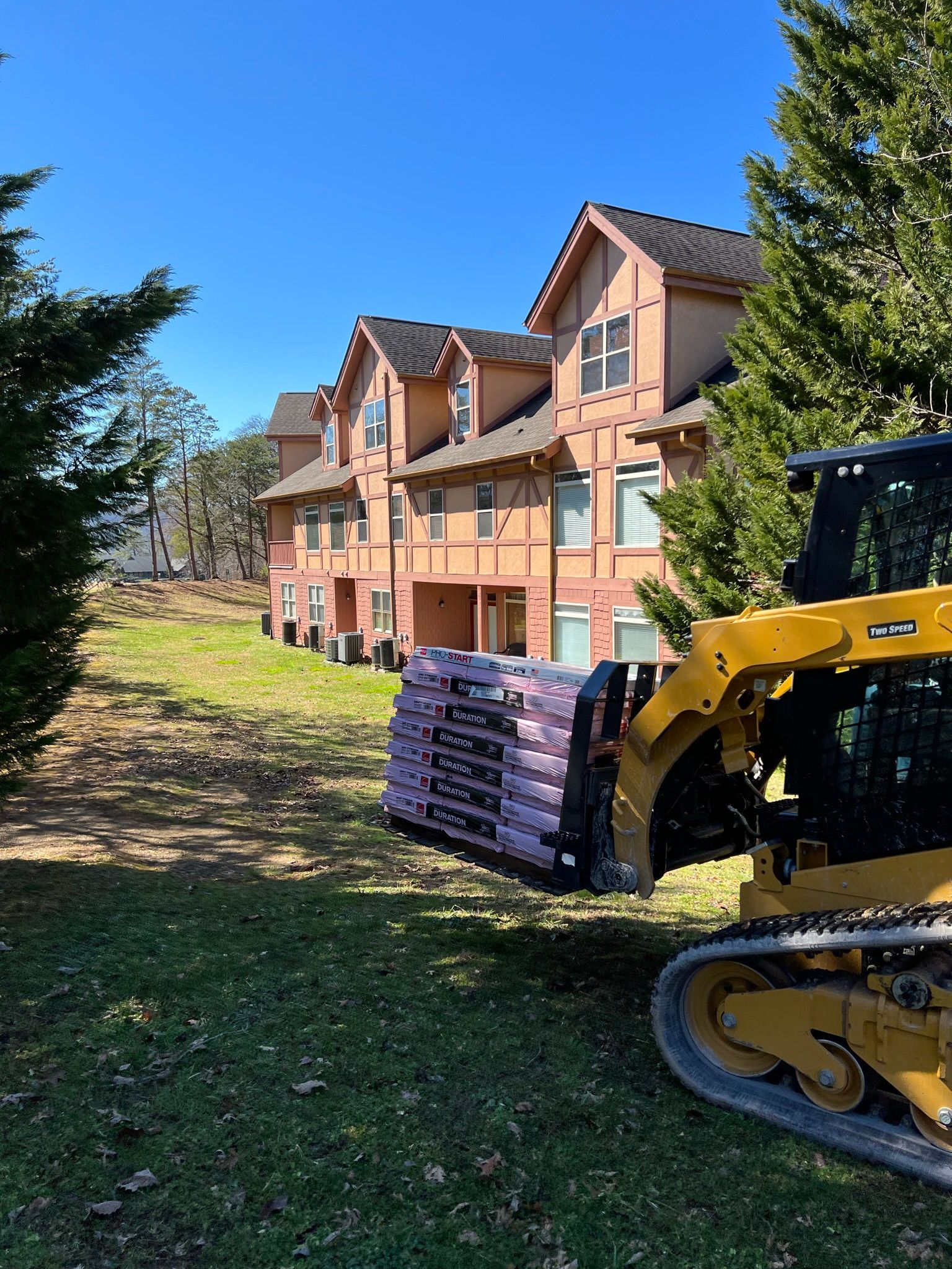 A yellow skid steer loader carries a pallet of pink insulation in front of a multi-story apartment building on a sunny day.