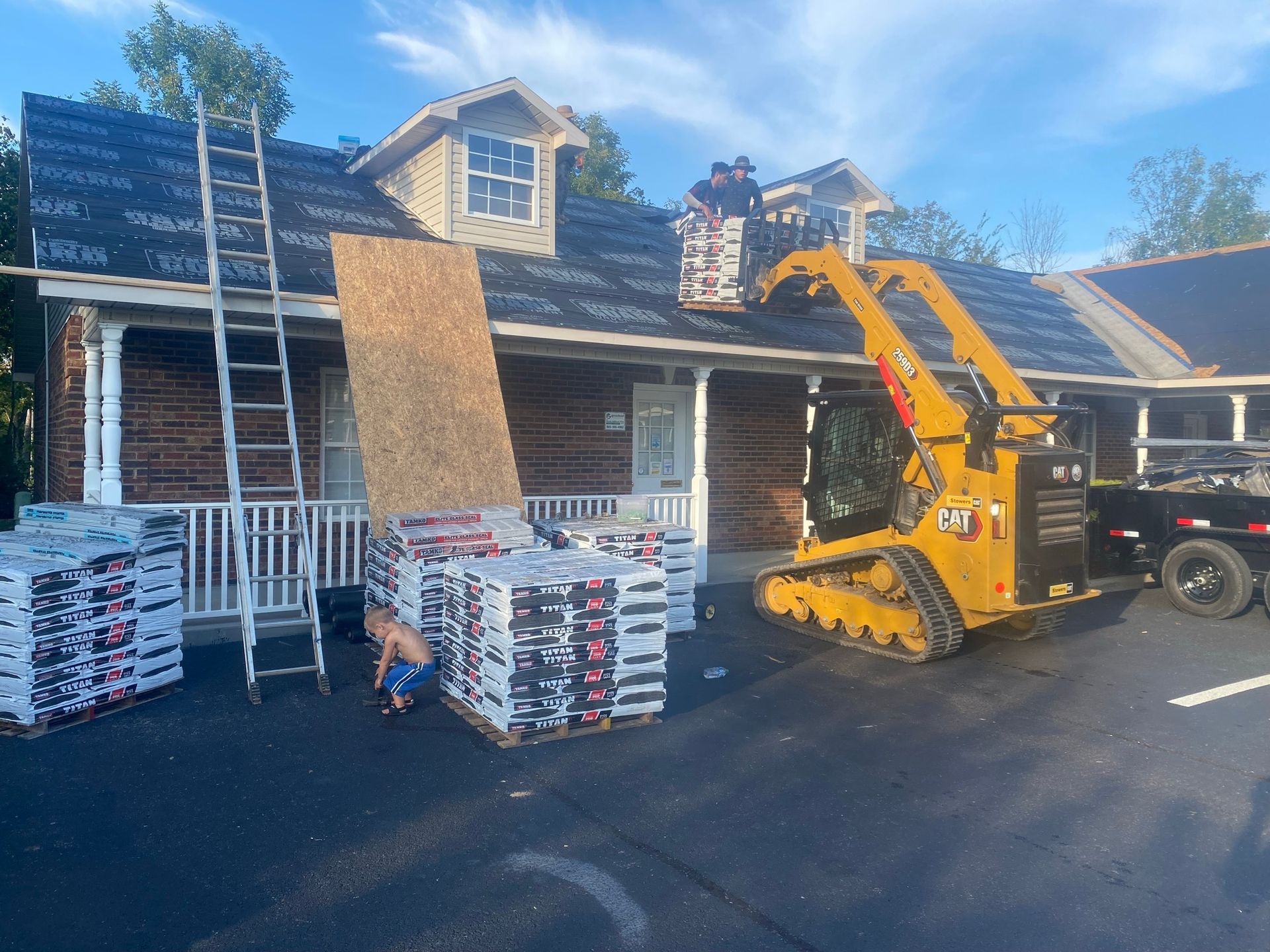 Workers use a yellow skid steer to lift materials to a roof under construction, with pallets of shingles on the pavement.