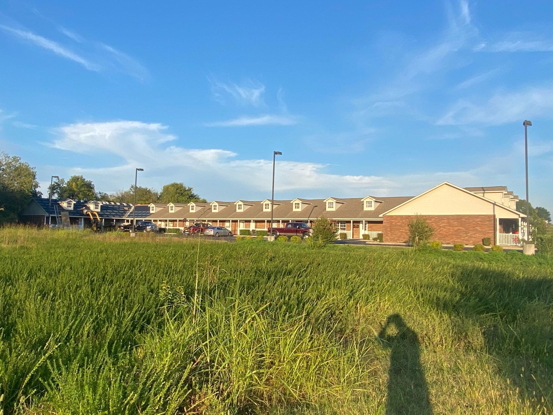 A long, low-slung building with a brick exterior stands behind a large, grassy field under a bright blue sky.
