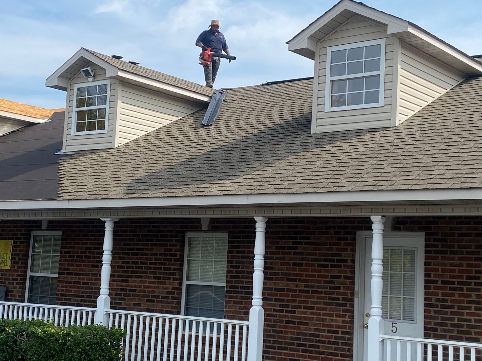A person standing on a brown shingled roof, using a leaf blower to clean debris from between two dormer windows.