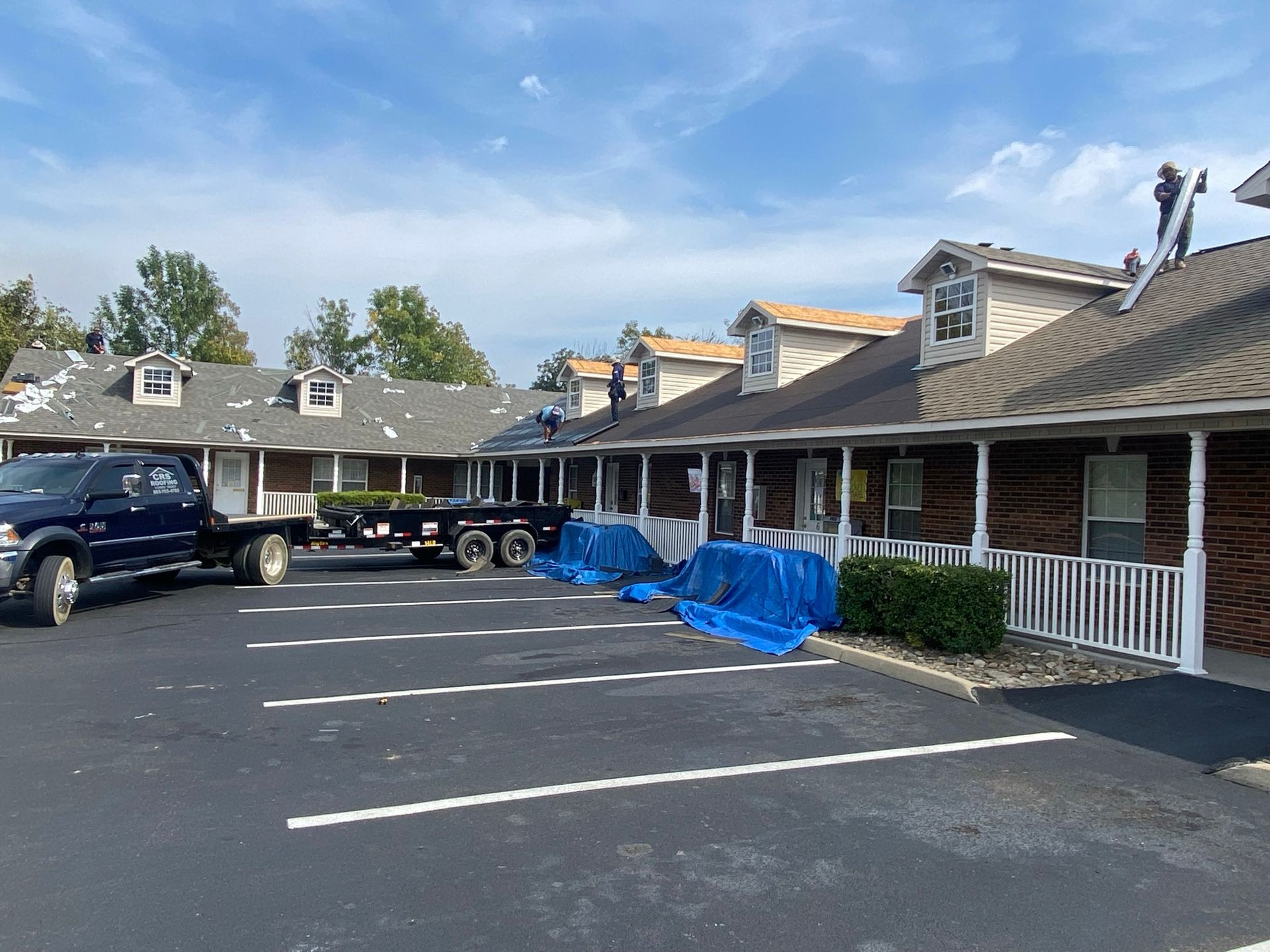 Roofers work on the shingled roofs of a long, brown building with white trim, with a truck and trailer in the lot.