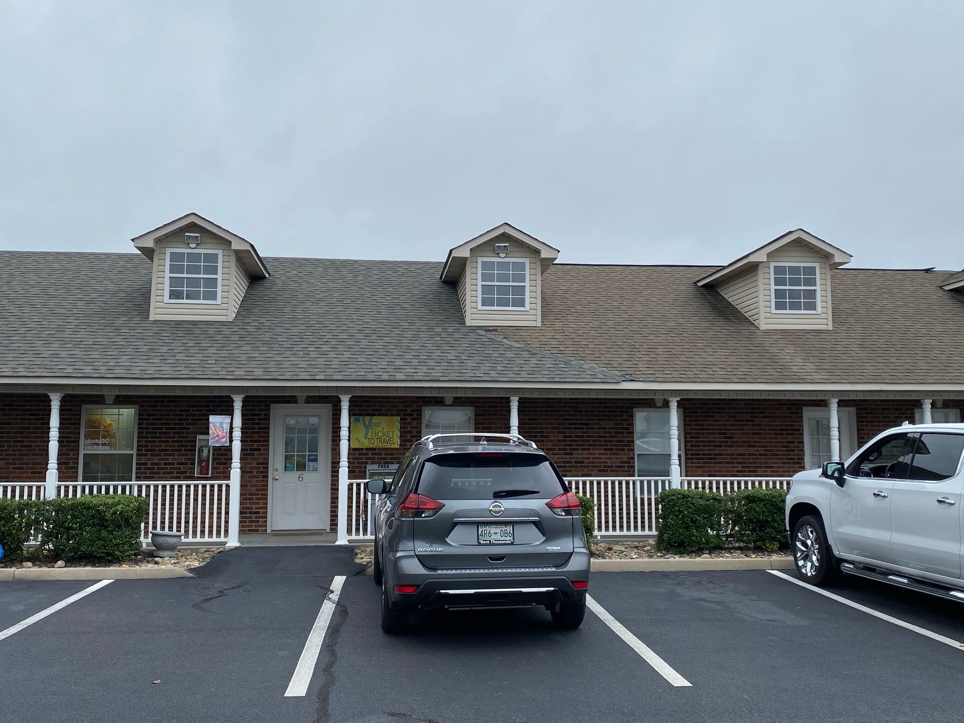 A brick office building with three dormer windows and a front porch, featuring two parked cars in the foreground.