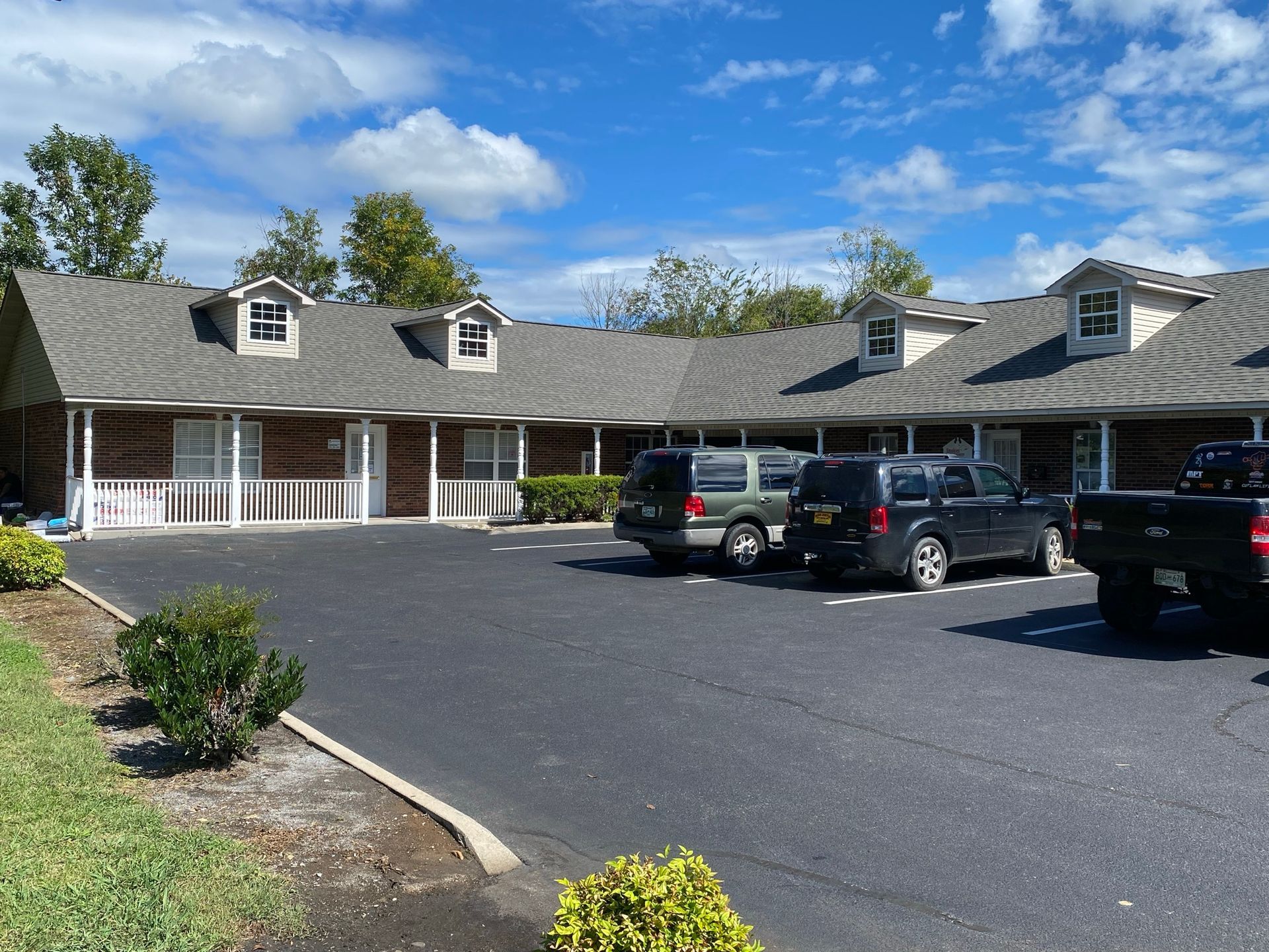 A one-story brick building with dormer windows and a dark parking lot featuring parked cars under a blue sky.