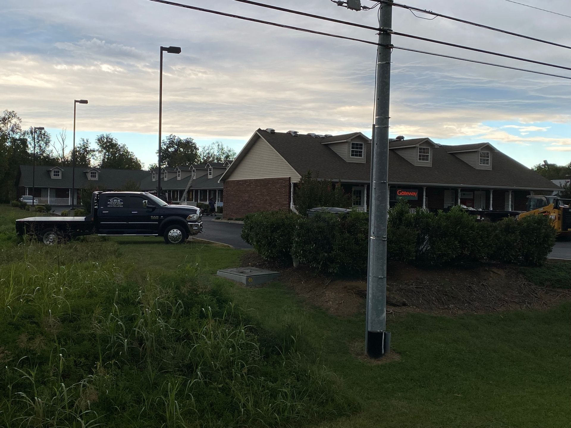 A low-angle view of a commercial strip mall with a dark pickup truck parked in front, bordered by green grass and shrubs.