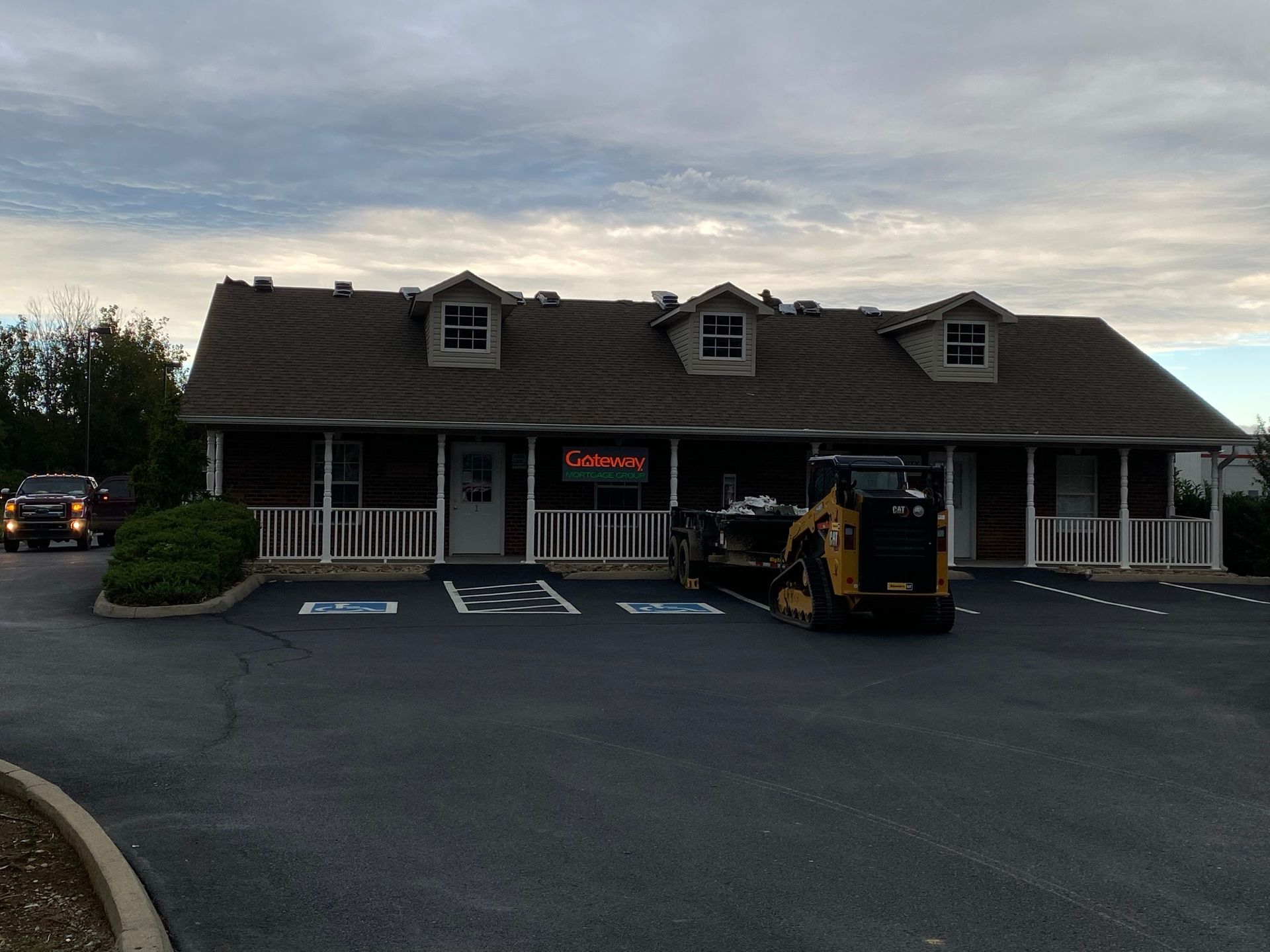 A single-story office building with a brown roof and a yellow skid-steer loader parked in the front parking lot.