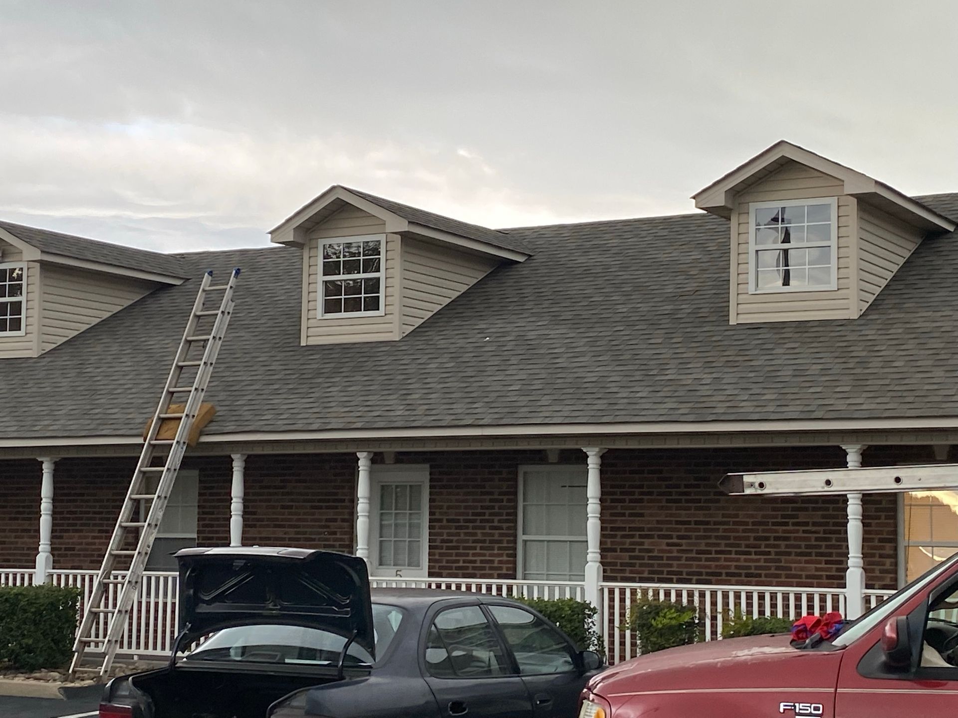 A ladder leans against the roof of a brick building featuring three dormer windows, with cars parked in the foreground.