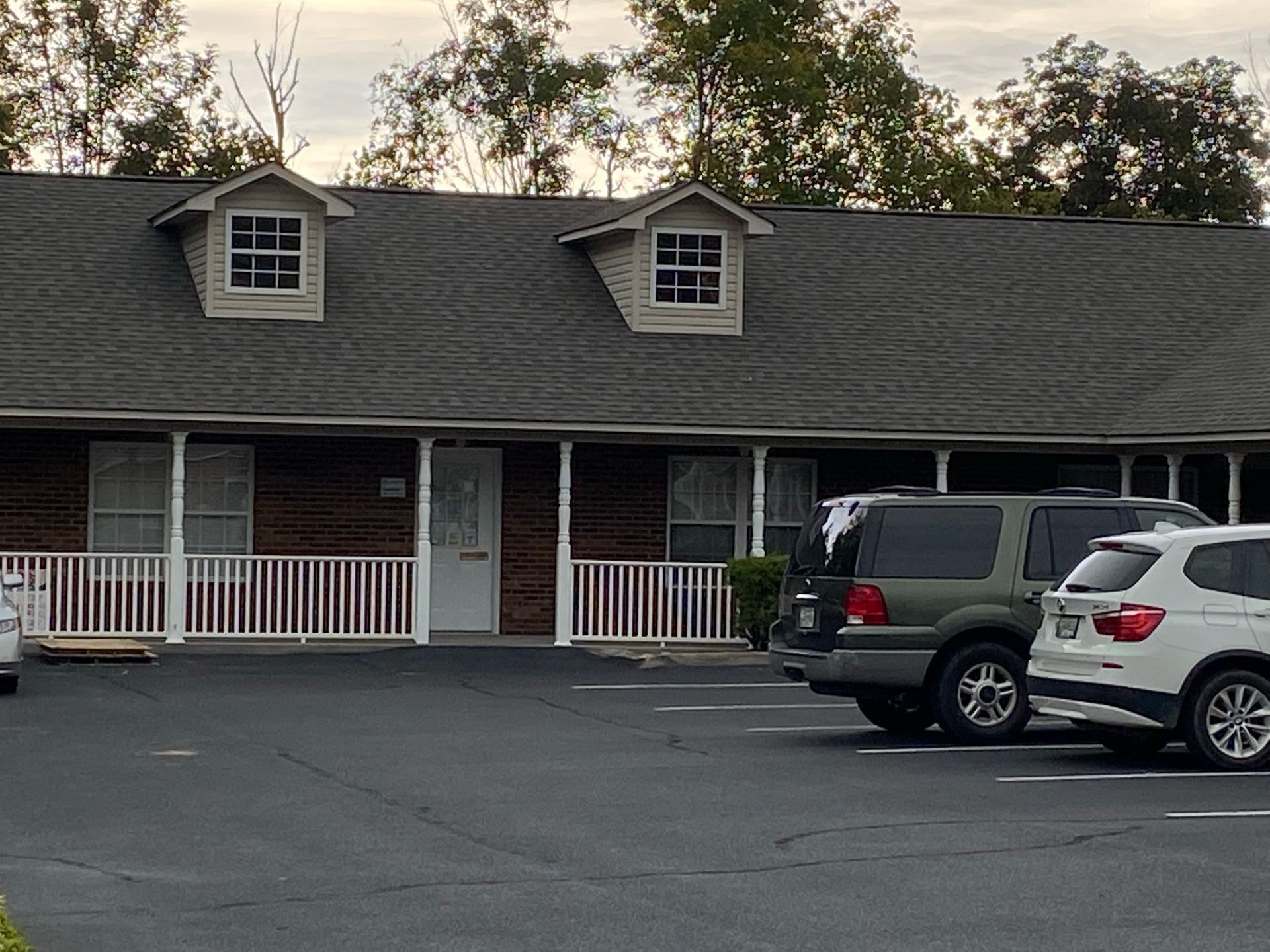 A brick building with white railings and two dormer windows, with a green SUV and a white SUV parked in front.