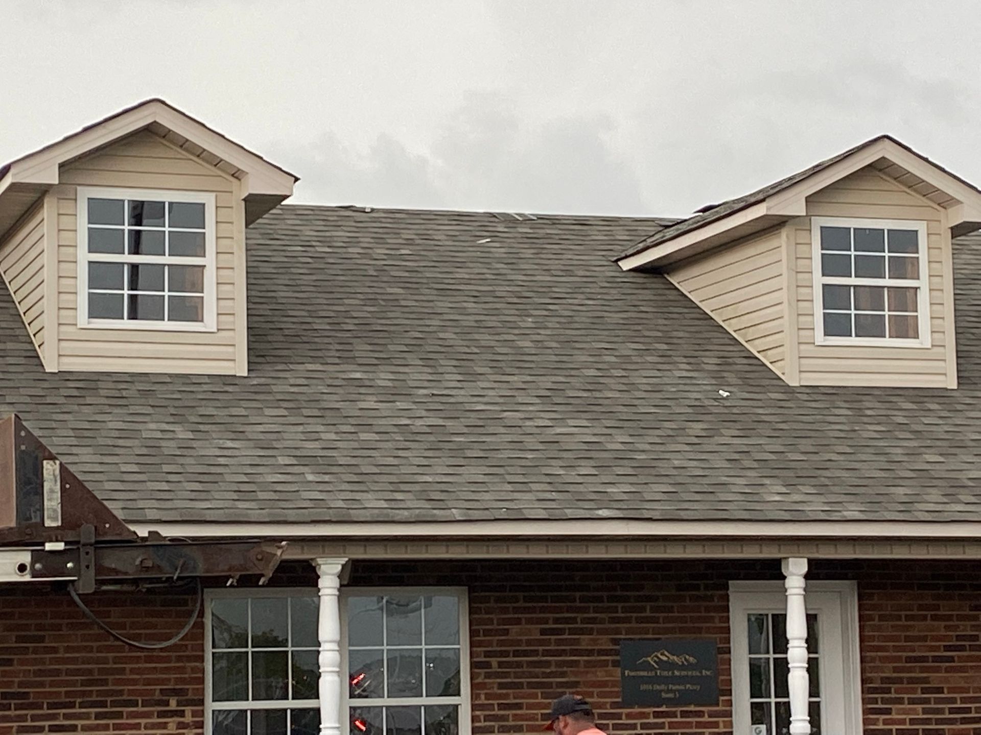 A brick building exterior featuring two tan dormer windows on a gray shingled roof under a cloudy sky.