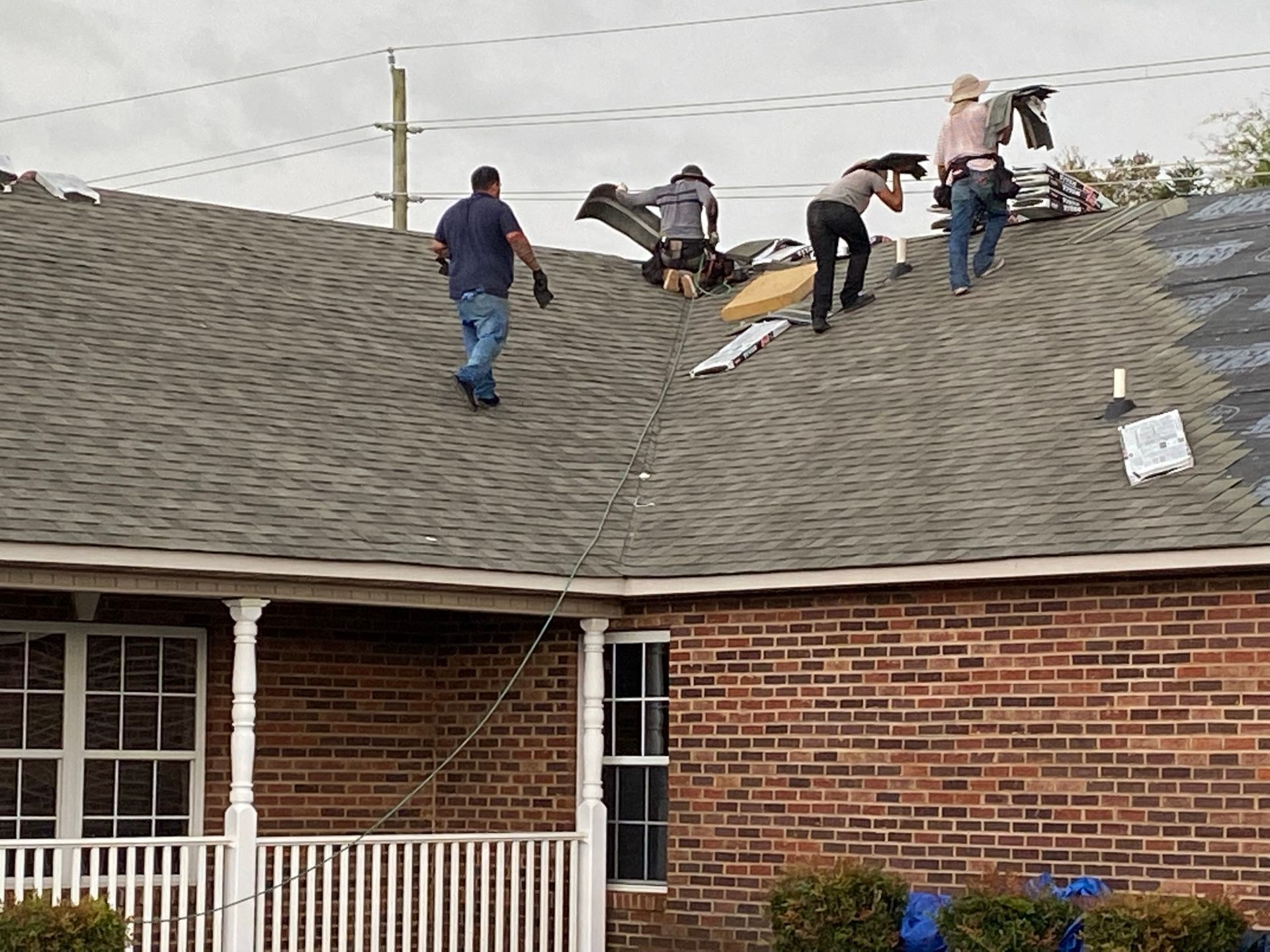 Four people in work clothes walk on a residential house roof, removing old shingles and carrying materials.