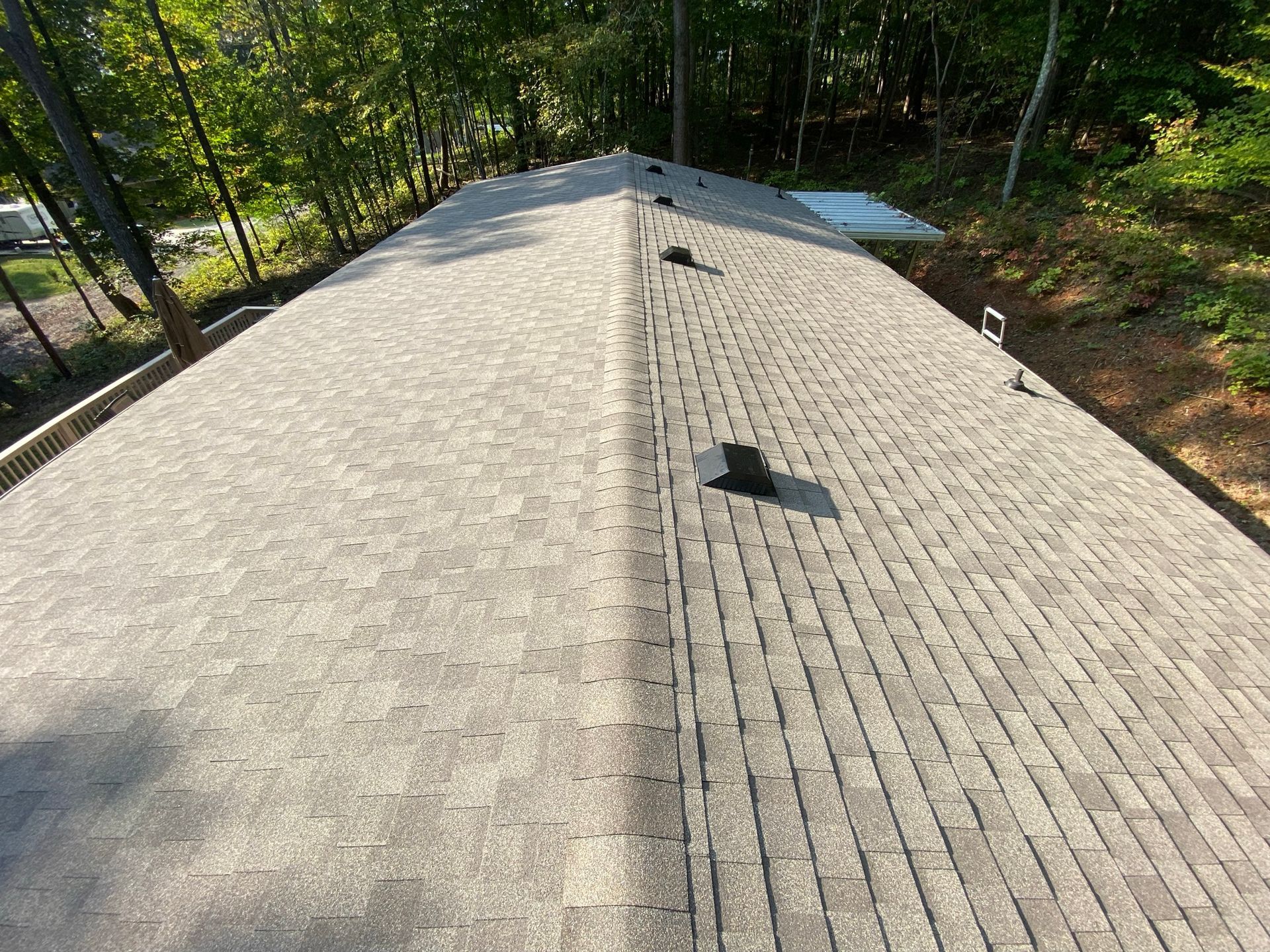 A high-angle view of a gray shingled roof with several black vents, set against a background of trees.