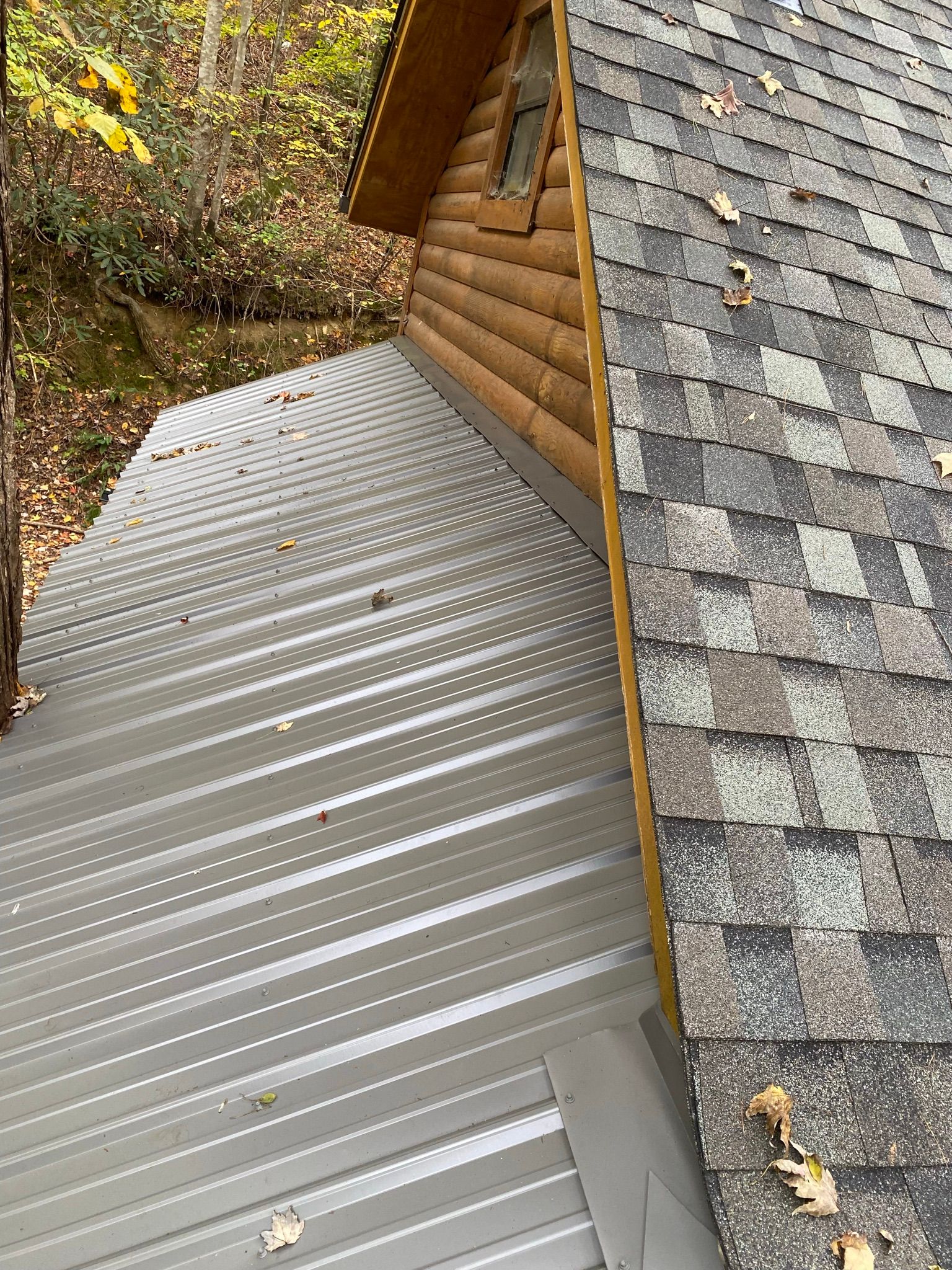 A metal roof meets a shingled roof and a log cabin wall in a wooded setting, featuring a flashing transition.