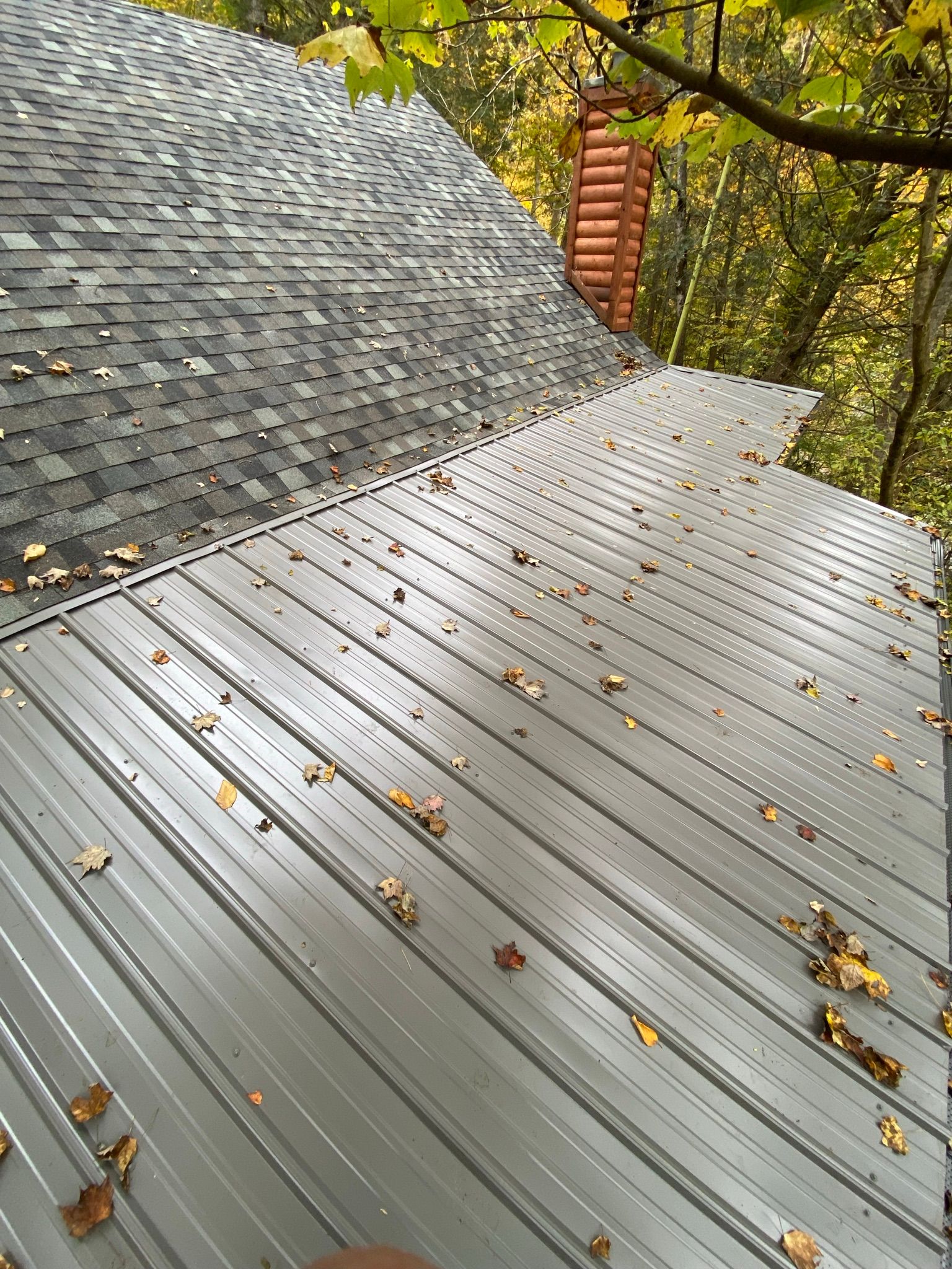 A gray, ribbed metal roof transition onto an asphalt shingle roof, with scattered autumn leaves and a brick chimney nearby.