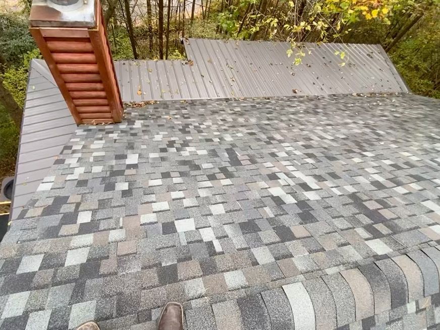 A high-angle view of a gray asphalt shingle roof featuring a brick chimney and metal roof sections.