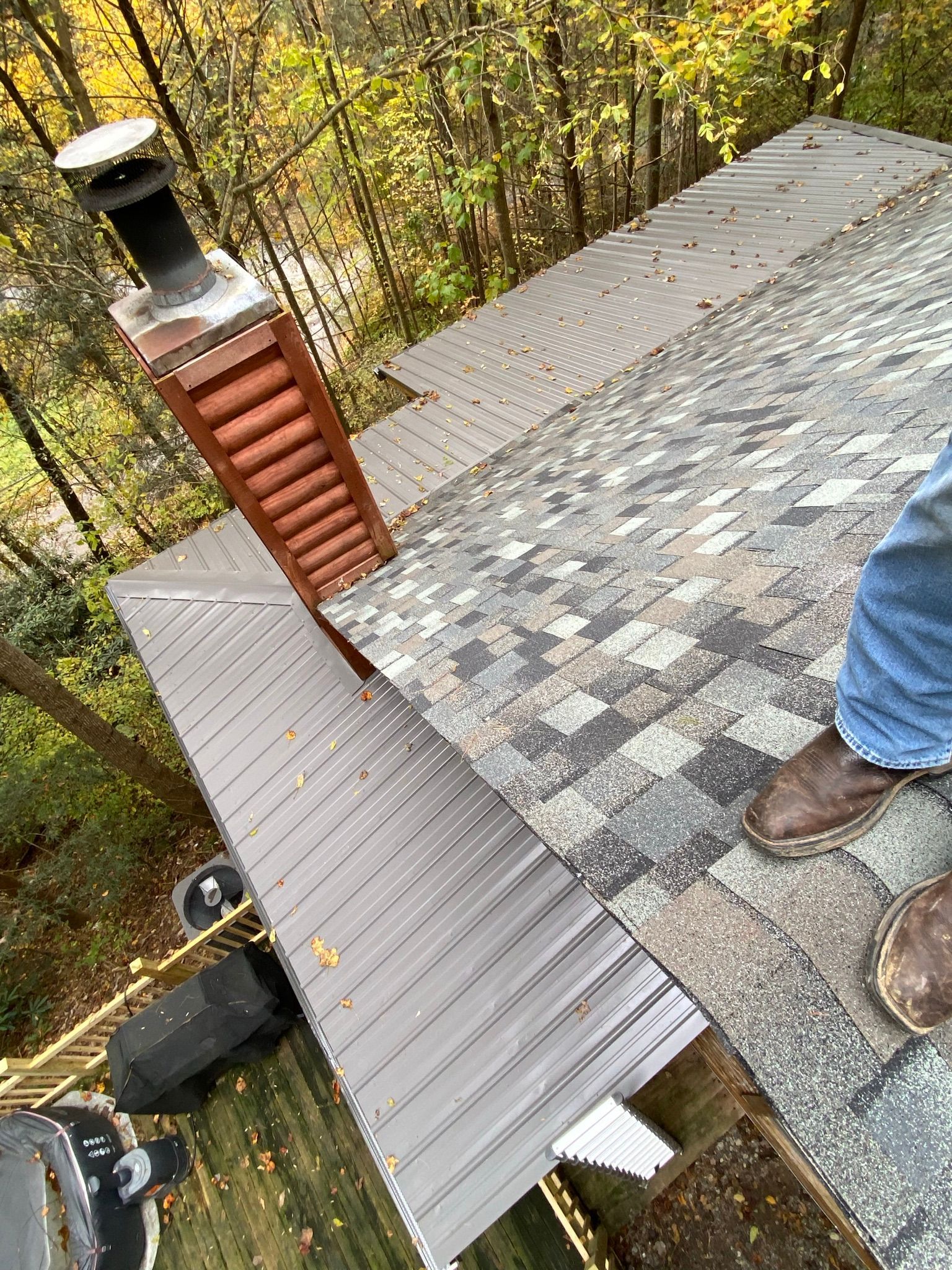 A view from a roof showing a brick chimney, gray metal roofing, asphalt shingles, and someone's legs in blue jeans.