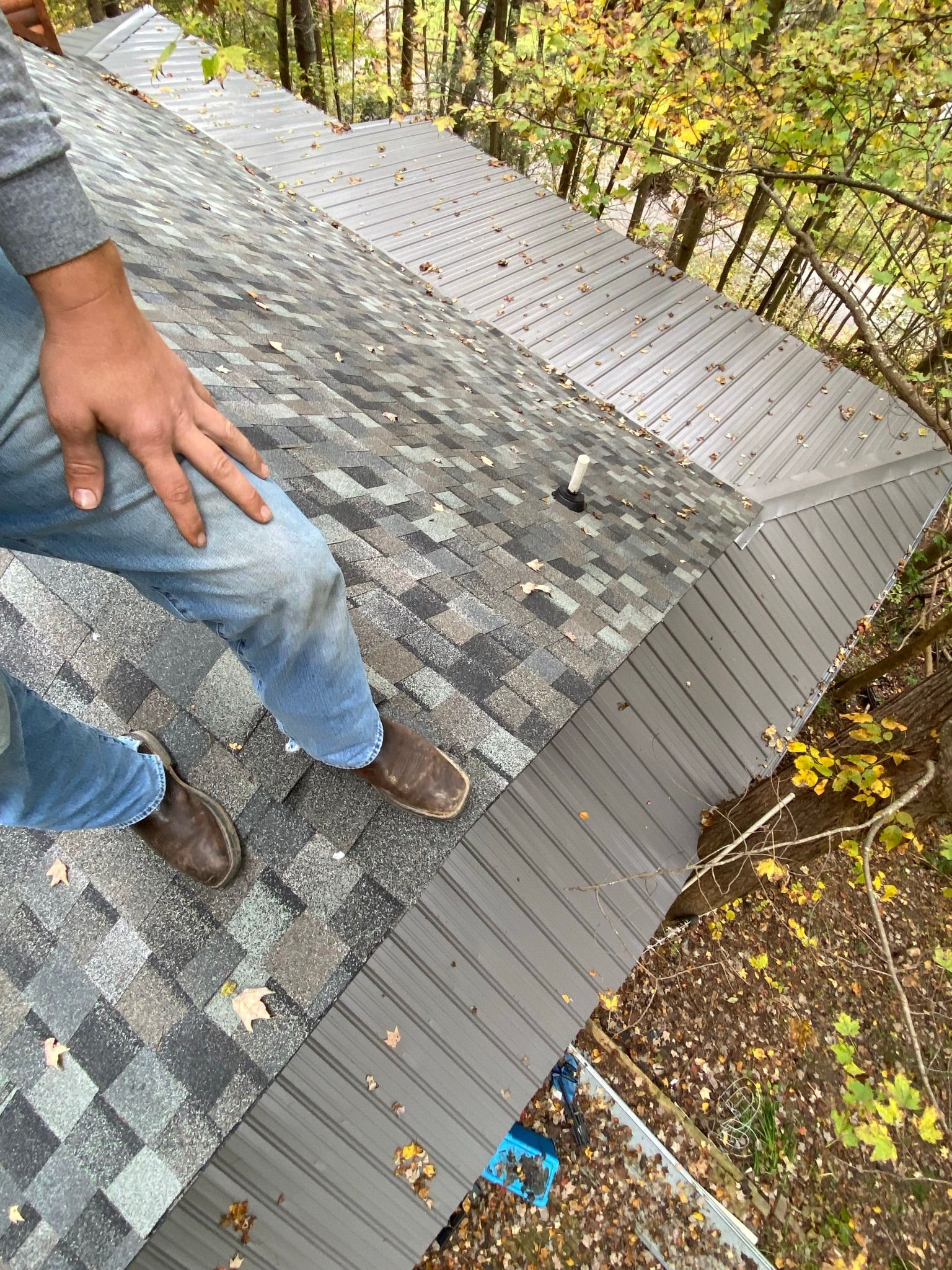 A person stands on a shingled roof next to metal flashing, with trees visible in the background.