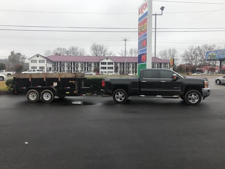 A black pickup truck towing a flatbed dump trailer parked in a gas station lot with a tall fuel sign in the background.