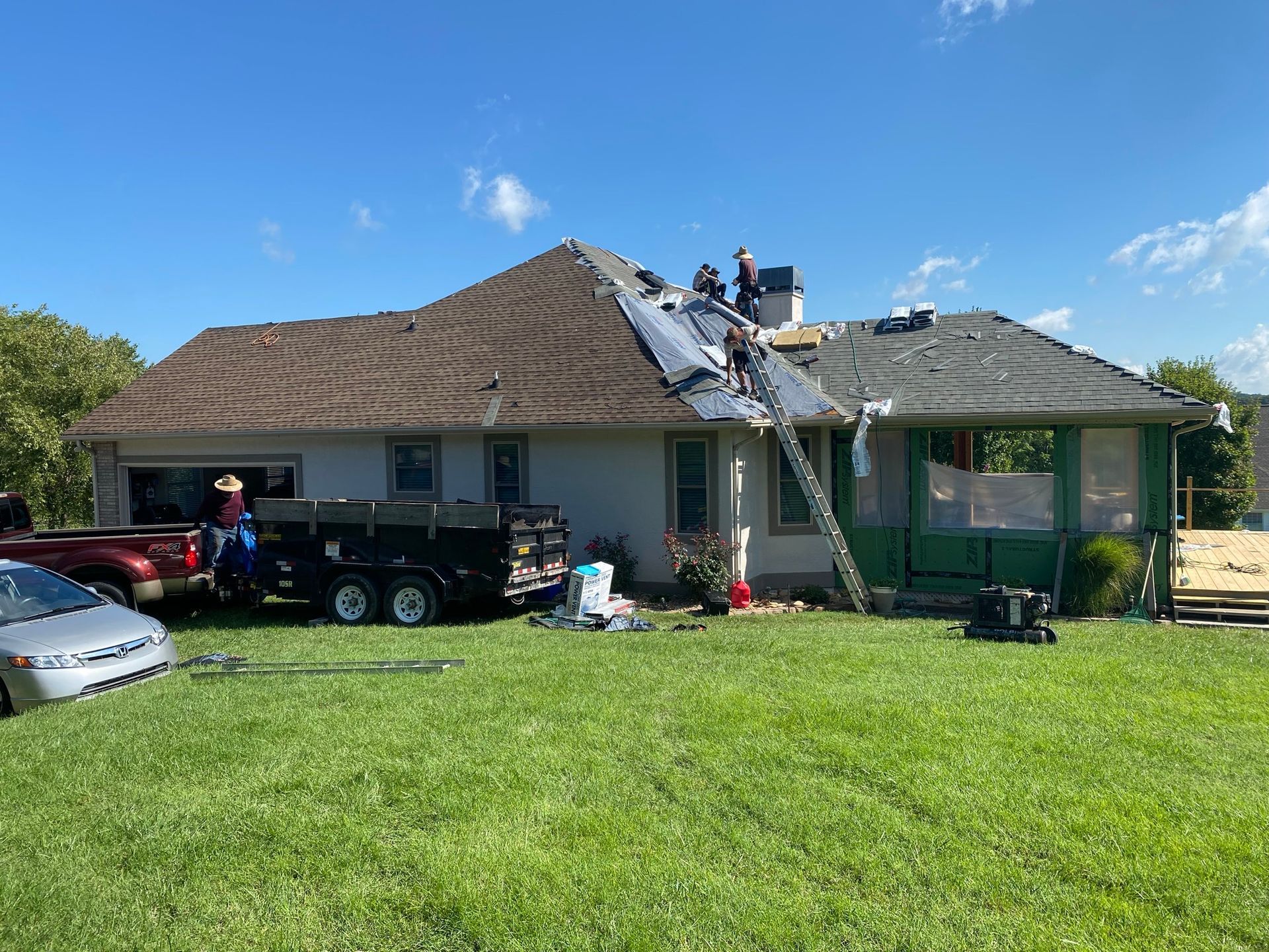 Workers repair the roof of a house on a sunny day, with a trailer parked in the yard.