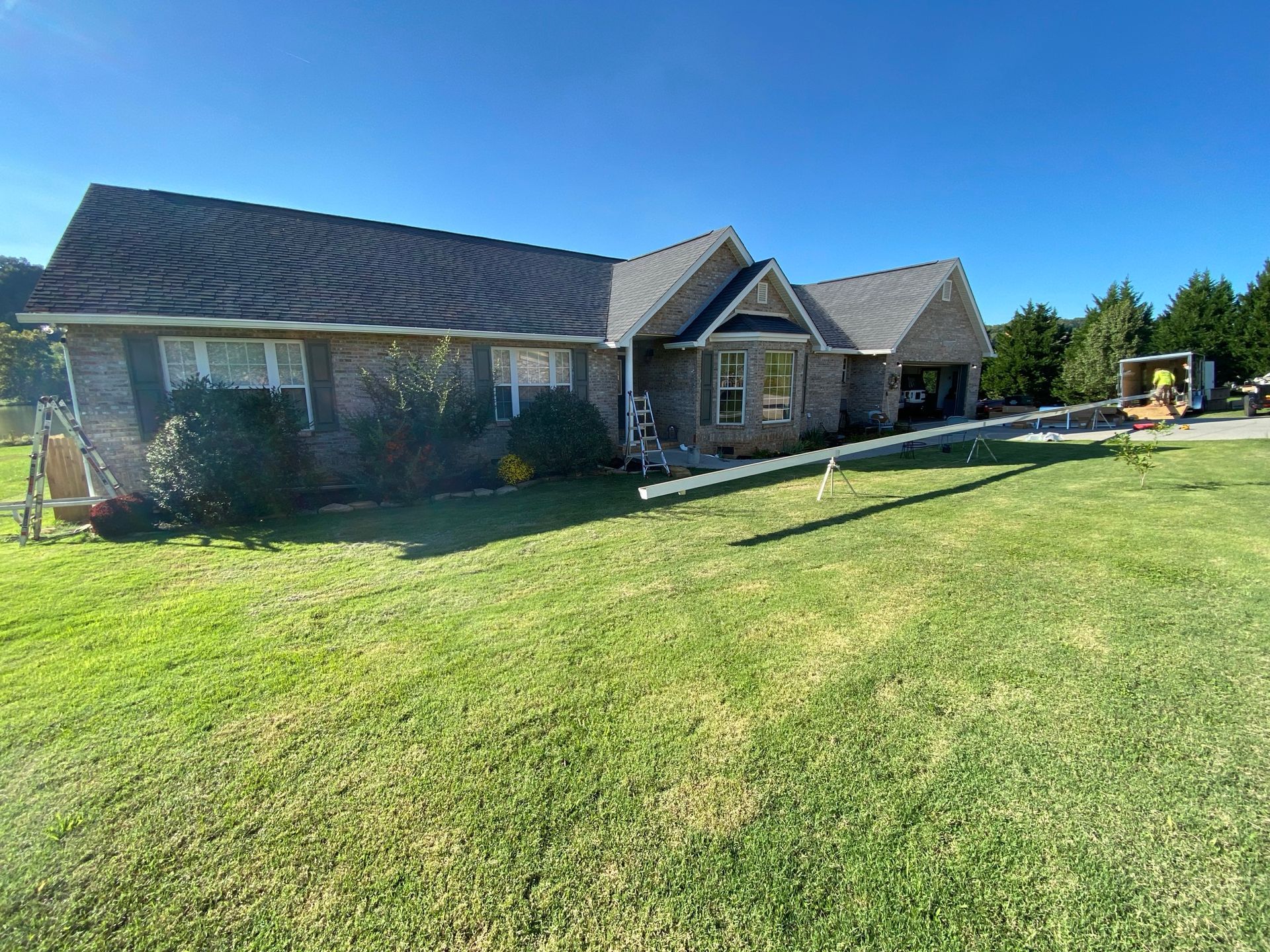 A brick house under a blue sky, with a long white gutter or pipe resting on a sawhorse in the grassy front yard.