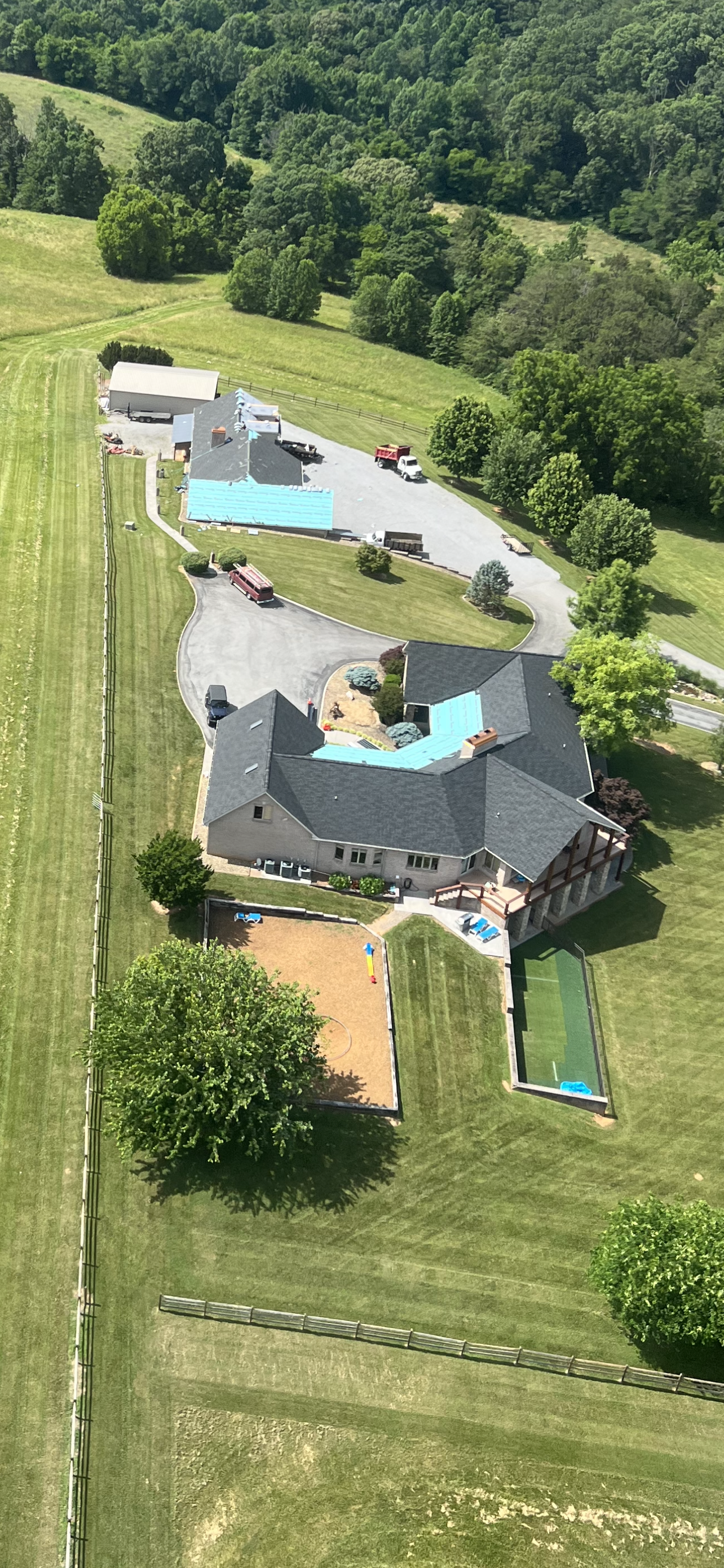 Aerial view of a sprawling house with a blue roof, a pool, and outbuildings surrounded by large green grassy fields.