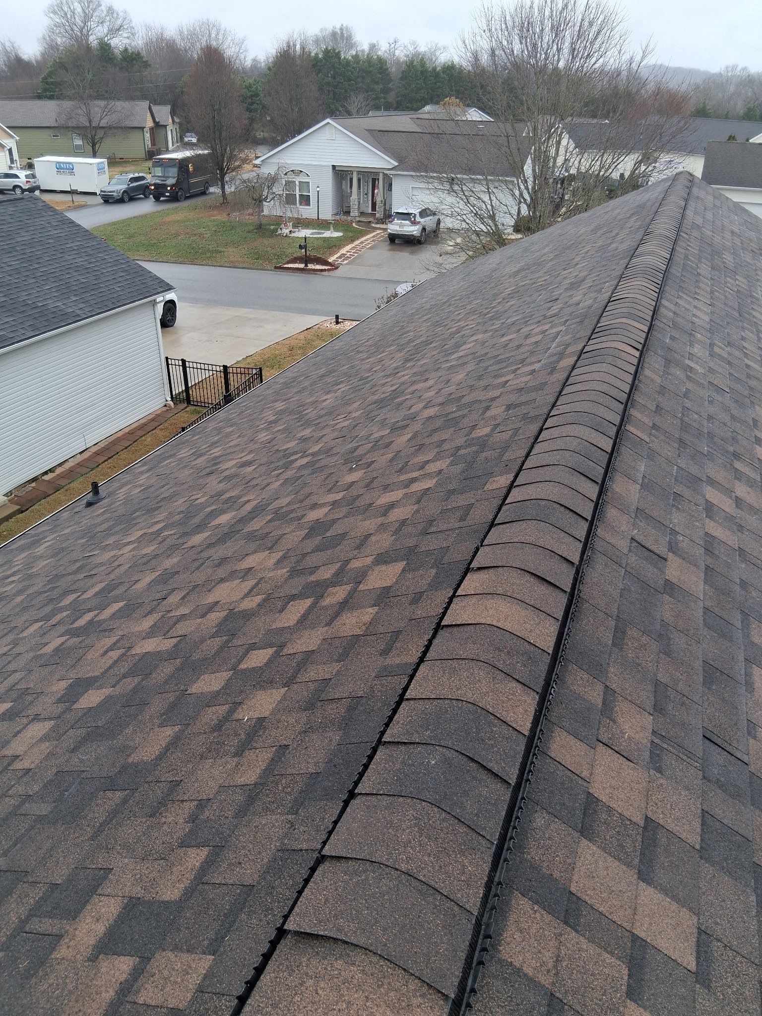 A high-angle view looking down the shingled peak of a residential roof, overlooking a driveway and neighborhood street.