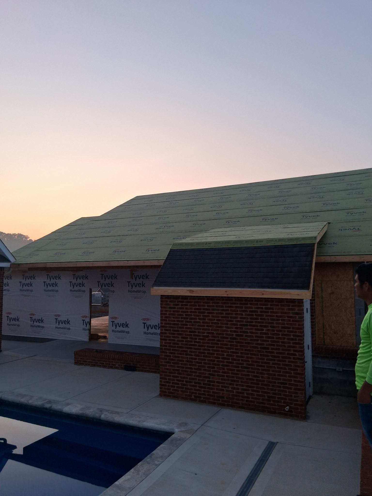 A residential building under construction next to a swimming pool, featuring a partially shingled roof at sunset.