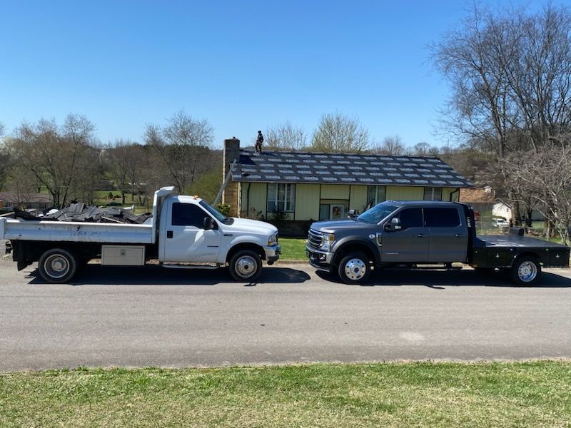 A white and a gray work truck parked in front of a house where a person is working on the roof under a blue sky.
