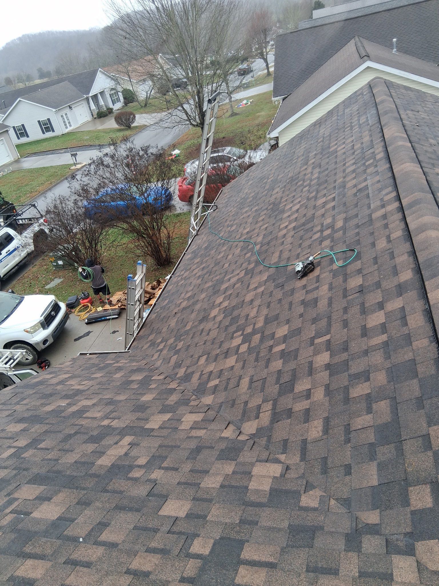 A view looking down a brown shingled roof towards a driveway, a parked white truck, and a ladder leaning against the house.