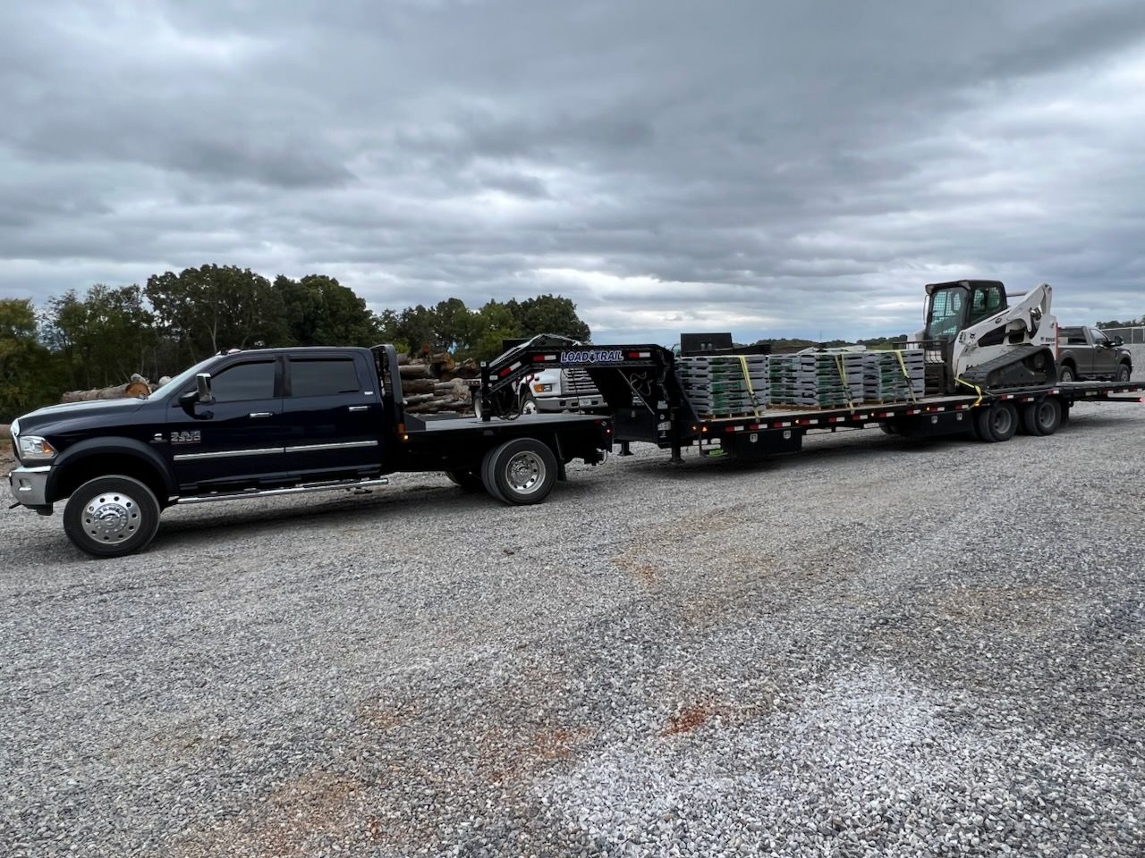 A black pickup truck with a flatbed trailer loaded with pallets and a skid-steer parked on a gravel lot under a cloudy sky.