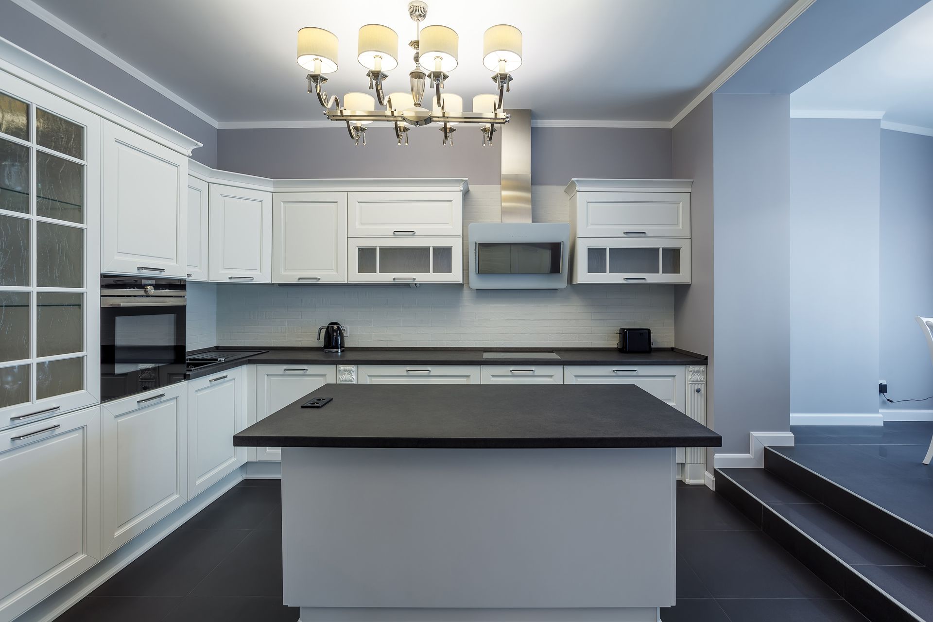 White kitchen with black countertops, island, and appliances. Chandelier hangs above.