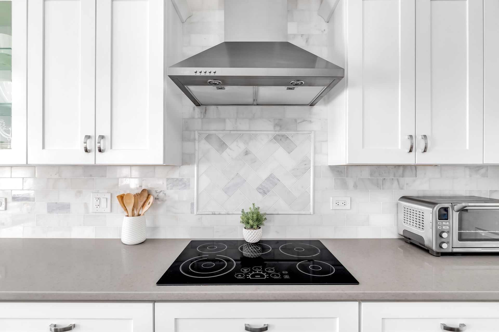 Kitchen with white cabinets, stainless steel range hood, black cooktop, gray countertop, and white backsplash.