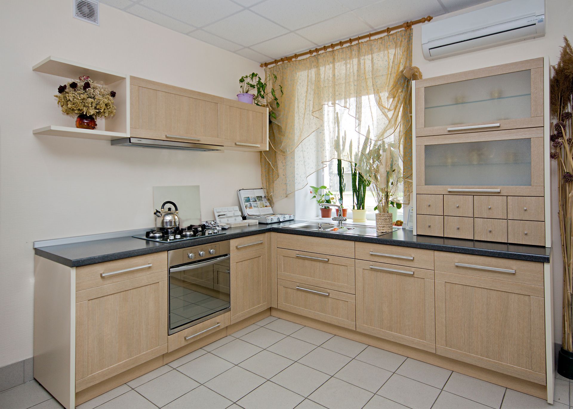L-shaped kitchen with light wood cabinets, gray countertops, and a window with sheer curtains.