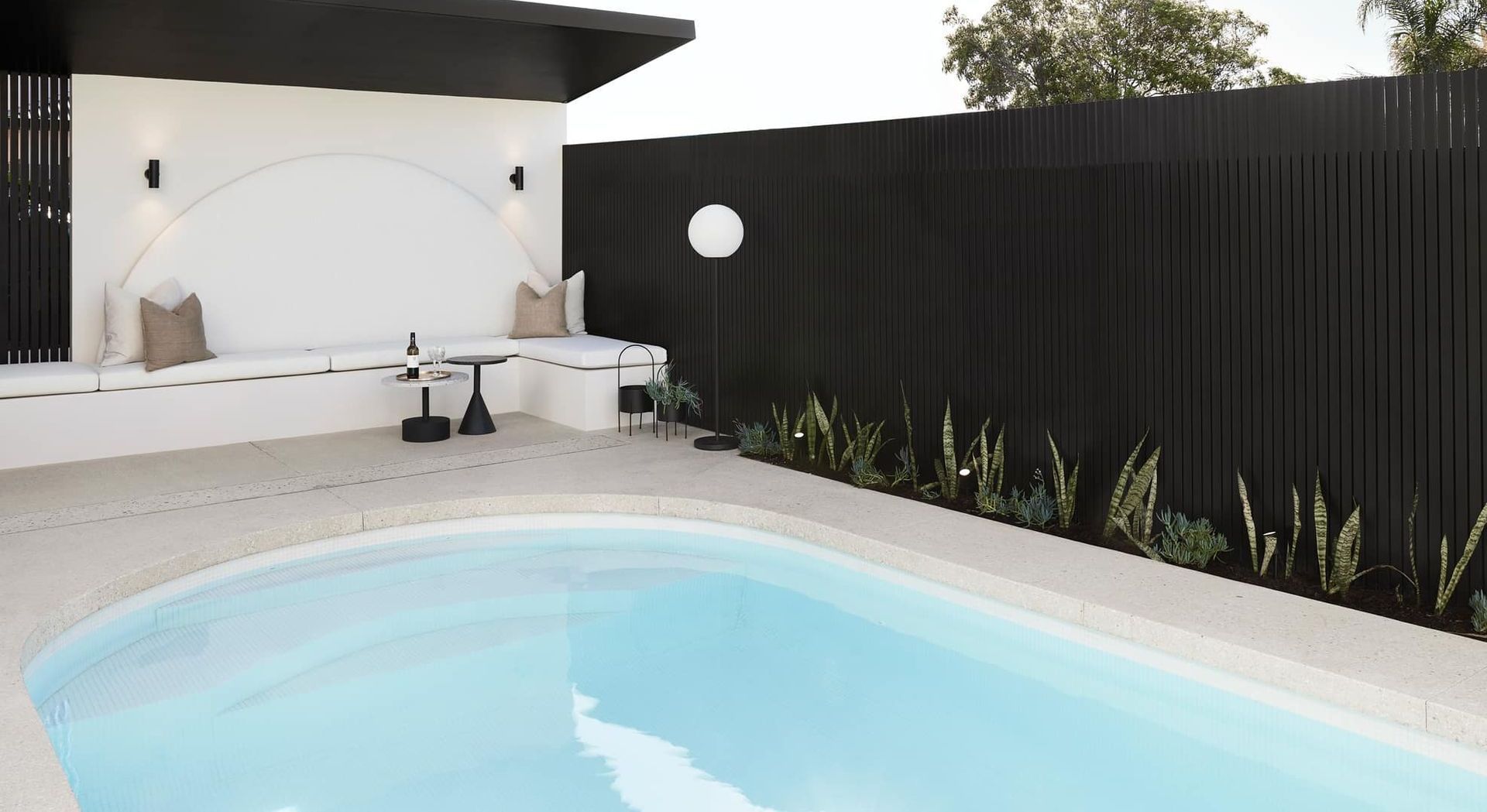 Backyard with a light blue pool, a white bench with pillows, and a black fence.